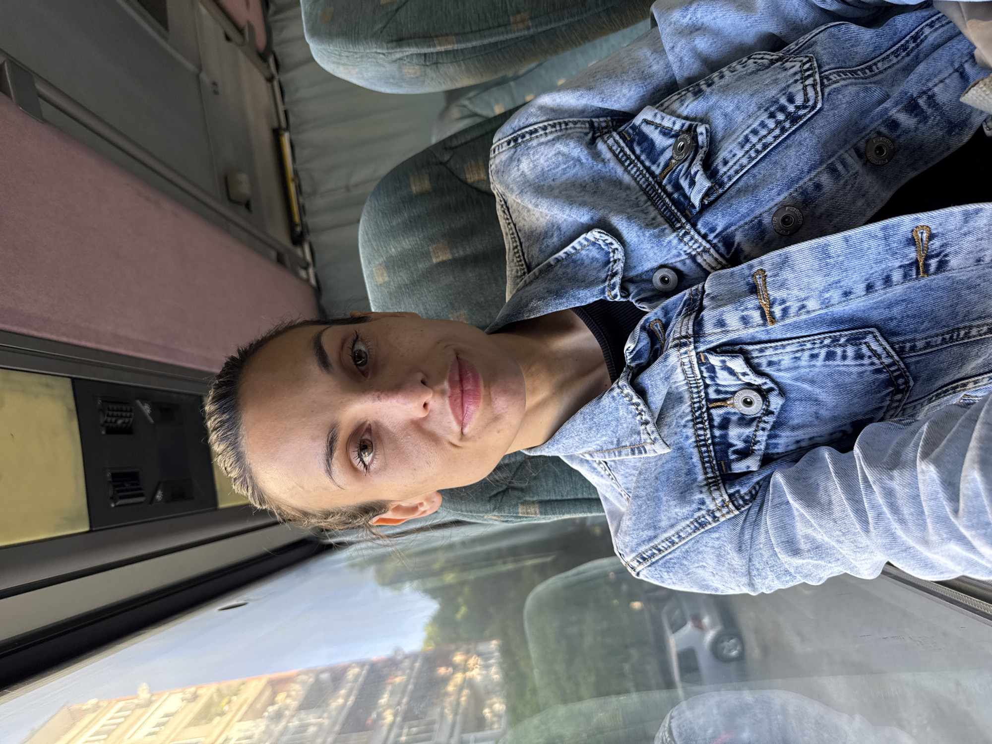 A woman is sitting on a vehicle, possibly a bus or train, with a window view of the city of Pleven, Bulgaria. The woman is wearing a denim jacket over a black top. The vehicle's interior features green patterned seats and a control panel. The window shows an urban landscape with buildings and a car in the reflection.