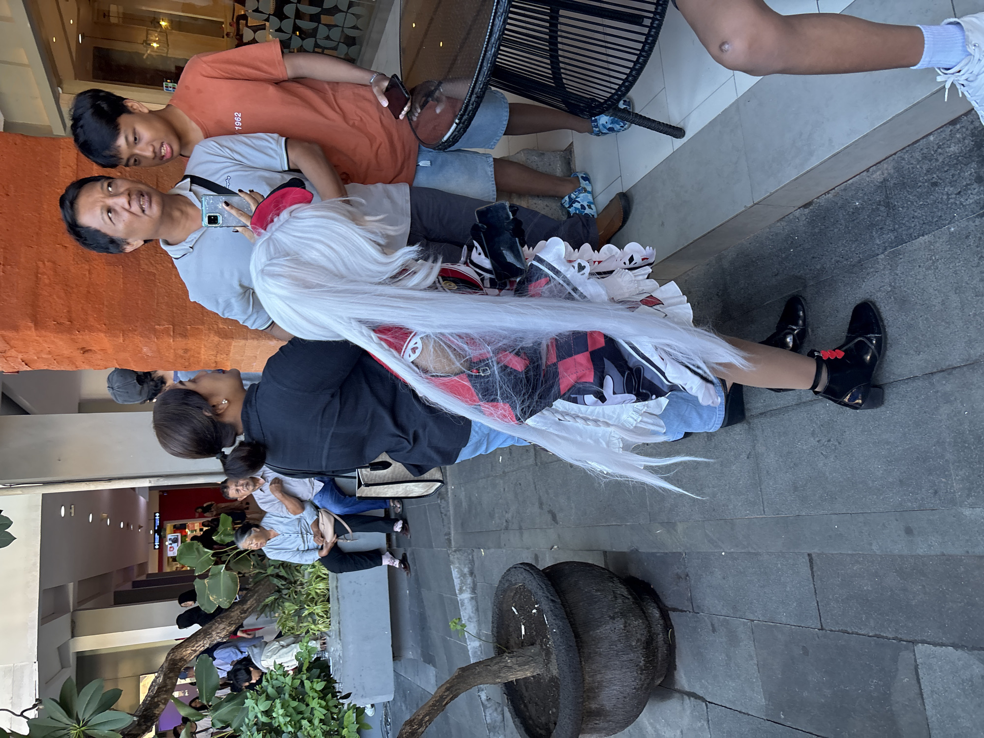 In Kuta, Indonesia, a group of people are gathered outdoors on a paved area in front of a building with orange brick accents. In the foreground, a person in a cosplay outfit with a long white wig and a red and black checkered dress is partially visible. To the right of the cosplayer, two men are standing. The man on the left, wearing a light gray polo shirt, is holding a smartphone. The man on the right, wearing an orange t-shirt with "1962" printed on it, is looking towards the cosplayer. Further in the background, several other individuals are seated and standing, some near planters with lush green foliage. A black wicker table with a round top is situated to the right of the group. The overall scene appears to be a public outdoor space, possibly near a commercial establishment.