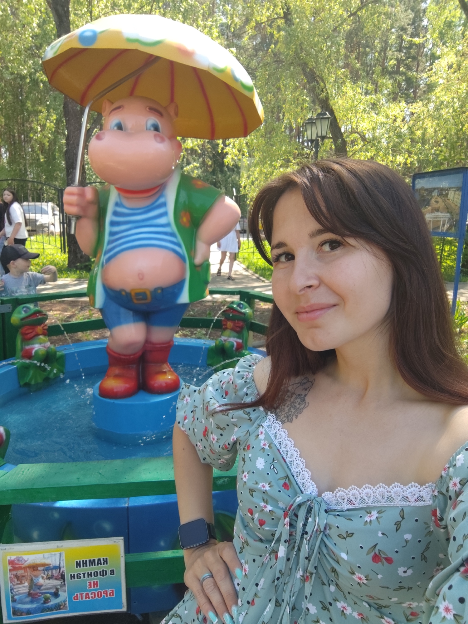 A young woman poses for a selfie with a cartoon hippo fountain in Chaikovsky, Russia. The hippo, wearing a striped shirt, green jacket, blue shorts, and red boots, stands in the center of the fountain holding a yellow umbrella. The woman wears a floral dress, a smartwatch, and a ring. A sign depicting the fountain and text in Russian is visible. In the background, other people and trees are present in the park setting.