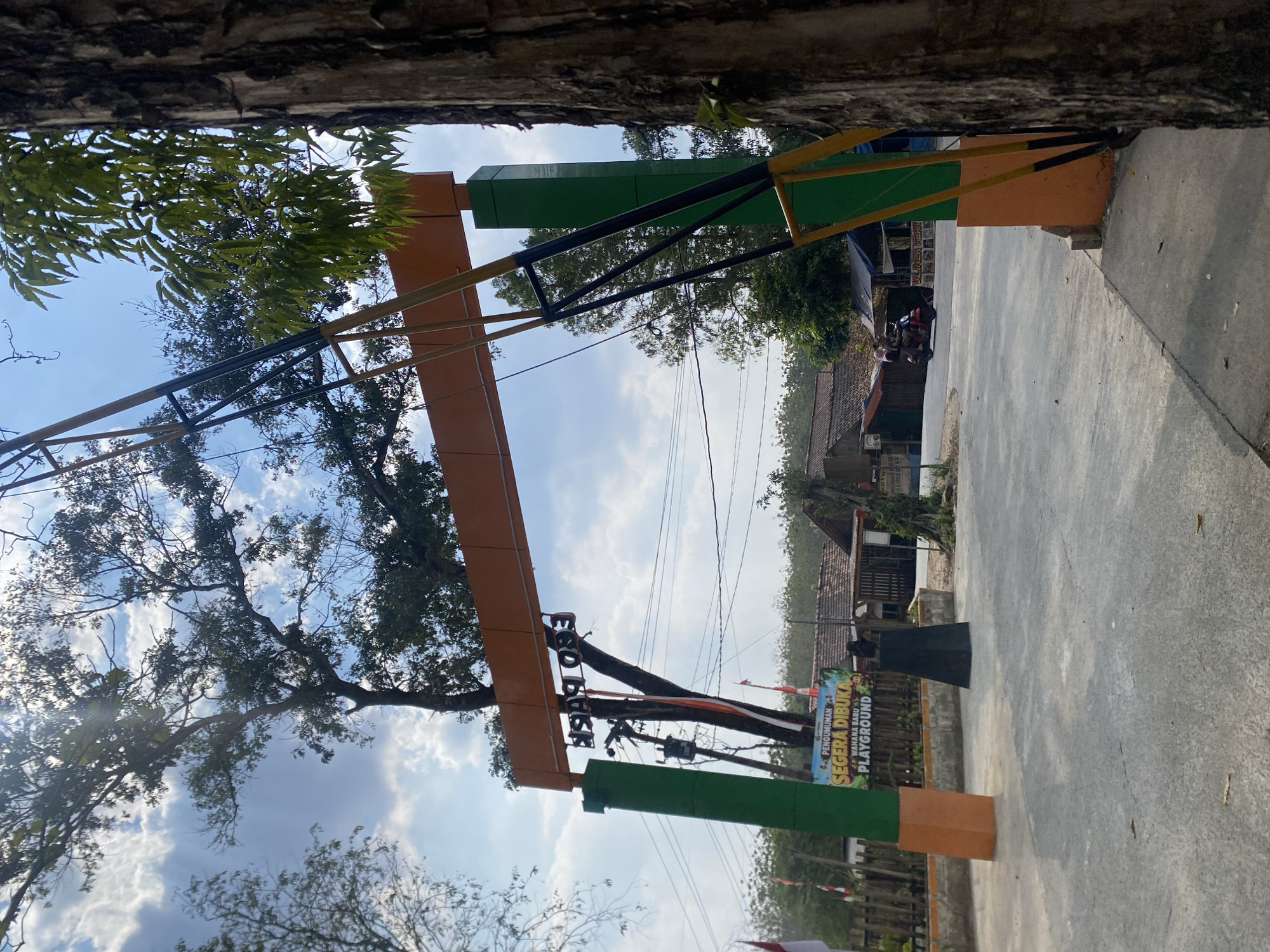 An eye-level, medium outdoor shot shows the entrance to "Eco Park" in Ketileng, Indonesia. The park entrance is an orange arch supported by green posts. The words "Eco Park" are written on the arch in black lettering, and are upside down. To the left of the entrance, a sign reads "PENGUMUMAN SEGERA DIBUKA WAHANA BARU PLAYGROUND" which translates to "ANNOUNCEMENT GRAND OPENING NEW ATTRACTION PLAYGROUND". Two red and white flags are visible to the left of the entrance. In the background, a red motorcycle with two passengers drives away from the entrance, past several small buildings. The ground is paved with concrete. Sunlight streams through the branches of a large tree on the right side of the frame.