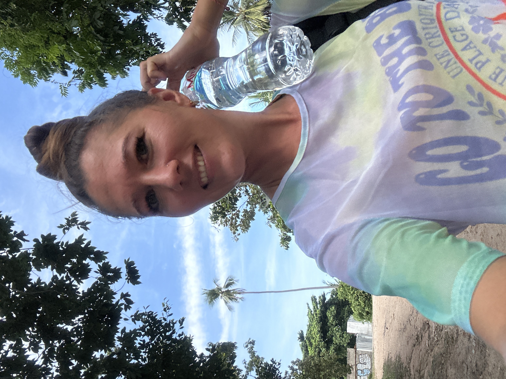A woman is taking a selfie outdoors. She has her hair pulled up in a bun and is holding a bottle of water to her ear. She is wearing a tie-dye t-shirt with the words "GO TO THE..." visible on it. In the background, there are palm trees and a cloudy blue sky. The location appears to be a beach or coastal area in Ko Samui District, Thailand. The ground is sandy and there are some structures in the distance that appear to be graffiti-covered walls or buildings.