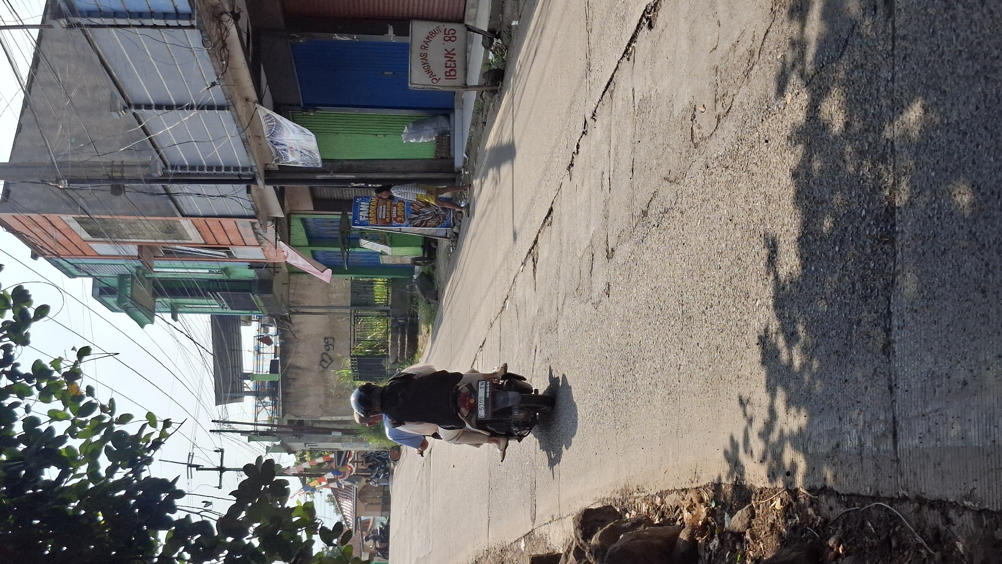 A motorcycle carrying two individuals travels down a paved road in Cilaku, Indonesia. The rider, wearing a dark helmet and a black backpack, is positioned in front, while a passenger in a light blue shirt and light-colored pants sits behind. The motorcycle's license plate reads "F 5399 XN". The road is lined with buildings, some of which have colorful facades in green, orange, and gray. Signs are visible, including one for "Pangkas Rambut Ibenk 85" and another advertising "FANI BAROKAH". A young woman in a striped shirt and yellow skirt stands near a food cart. In the background, power lines crisscross the sky, and some trees with green leaves cast shadows on the road. The environment appears to be an urban or semi-urban area with moderate sunlight.