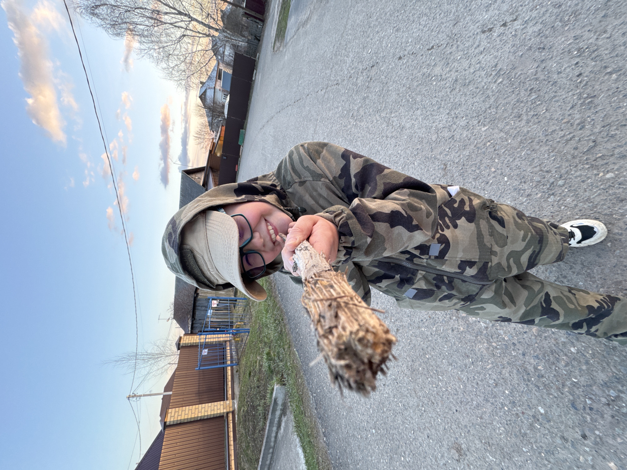 A young individual, likely a child or teenager, stands on a paved road in a residential area of Горный, Russia. They are dressed in a camouflage hooded suit and a light-colored bucket hat, wearing glasses and smiling brightly towards the camera. In their hands, they hold a rustic, broom-like object made from bound natural materials with a visible handle, extending it forward.
The scene takes place on an asphalt road, flanked by residential properties with various fences, including wooden and blue metal gates. Houses with brick and darker siding are visible in the background, along with bare trees, suggesting either early spring or late autumn. The sky is clear blue with scattered, wispy clouds displaying soft pink and orange hues, indicating either sunrise or sunset. The overall atmosphere conveyed by the person's expression is one of playfulness and happiness. The image is captured at a tilted angle, adding to its dynamic feel.