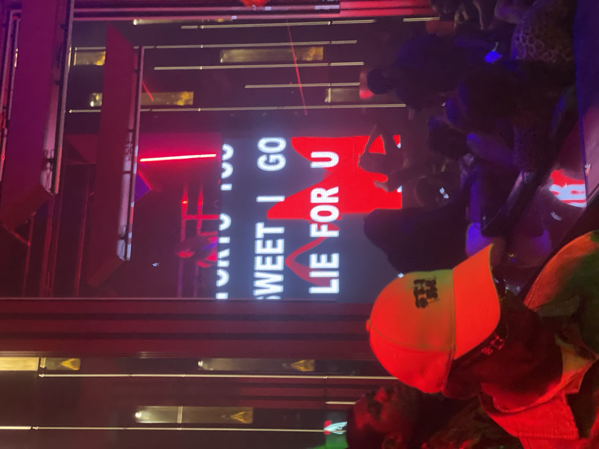 The image depicts a dimly lit, indoor venue, likely a nightclub or entertainment space, illuminated by vibrant red and pink neon lights. In the background, a large LED screen displays white text that reads "SWEET I GO LIE FOR U". The screen is partially obscured by a red, stylized graphic of a person's head and shoulders. The scene is populated by several individuals, mostly silhouetted against the bright lights. In the foreground, a person wearing a light yellow baseball cap with black lettering is the primary subject. Their face is cast in shadow due to the lighting, but they appear to be wearing glasses and a dark jacket. The cap has a logo that is partially visible. Other individuals are seated or standing around, with some interacting. The overall atmosphere suggests a lively social gathering or event, possibly in Abuja, Nigeria, given the context provided.