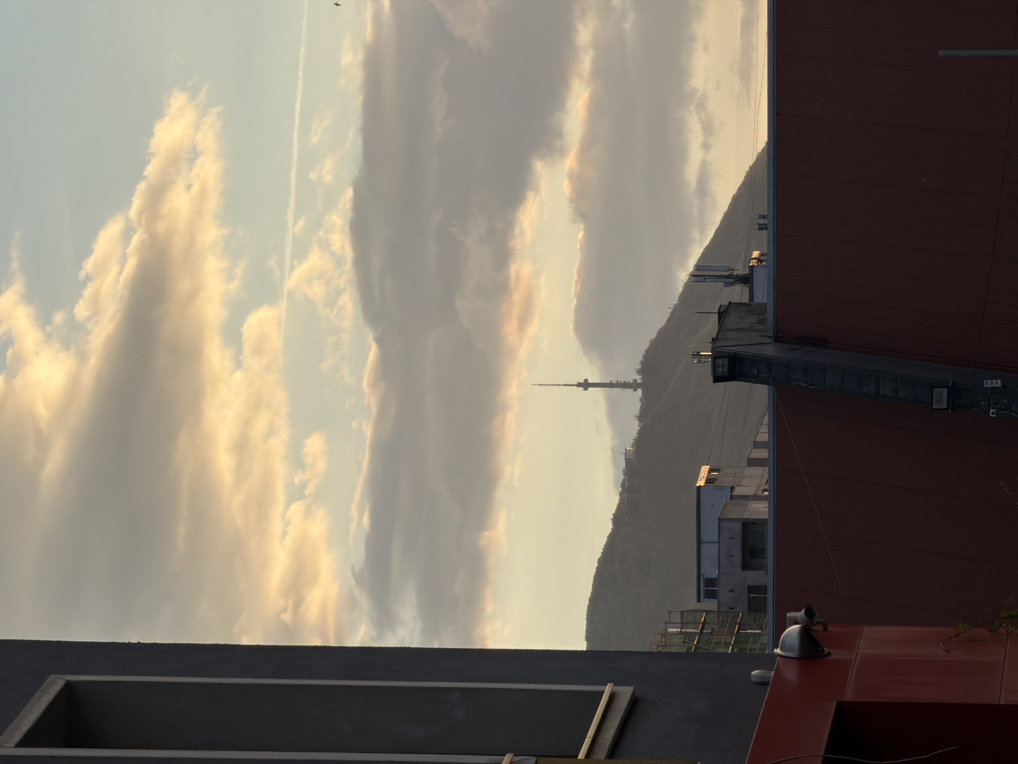 The image shows a view from a rooftop in Sofia, Bulgaria, facing towards the Kopitoto area of Vitosha Mountain. In the distance, the Vitosha TV Tower is visible on top of the mountain. The sky is filled with clouds, illuminated by the setting sun. In the foreground, there are elements of the rooftop, including a rectangular structure, a red surface with what appears to be a helmet, and parts of a dark red building. There is also construction work visible on the opposite side of the adjacent building. A contrail from an aircraft is visible in the sky.
