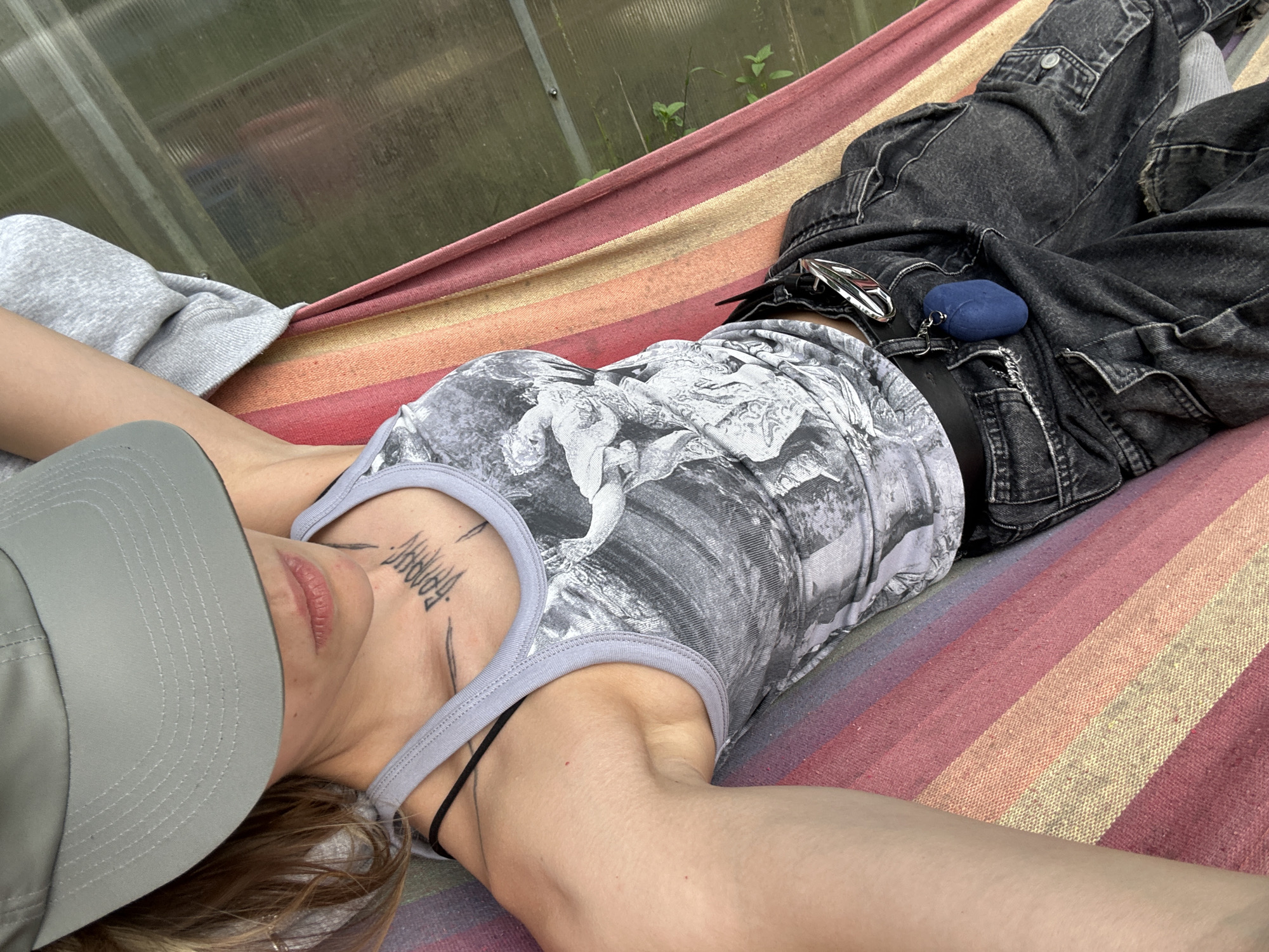 A person is lying in a hammock with a colorful striped pattern. They are wearing a grey t-shirt with a black and white graphic print, dark grey denim pants, and a light grey baseball cap. A blue earbud case is attached to their belt loop with a keychain. The background appears to be a greenhouse with corrugated plastic walls, showing some greenery outside. The image was taken in the city of Kozino, Russia.