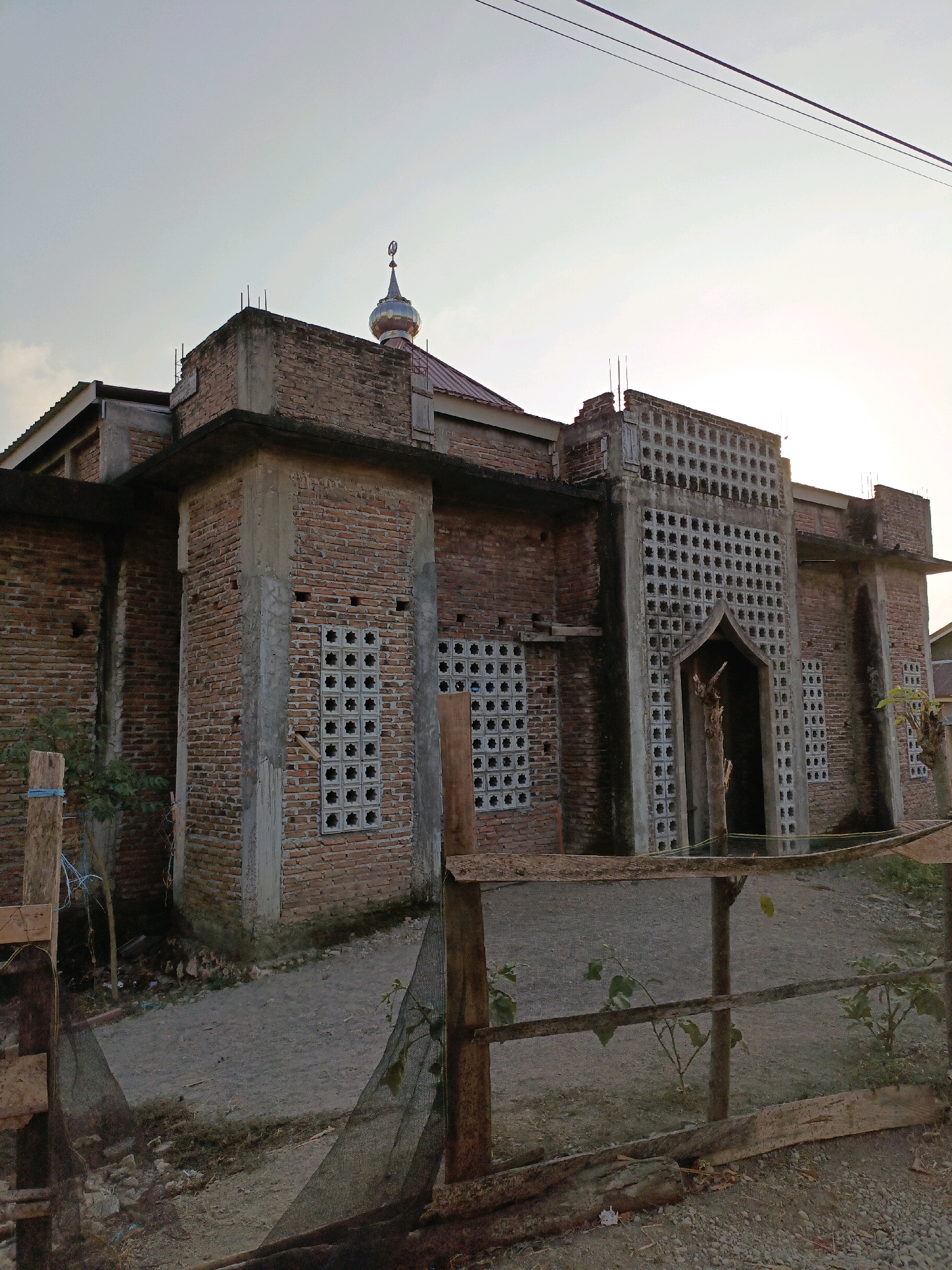 The image depicts a mosque under construction in Bulukumba, Indonesia. The building is primarily constructed of brick, with a concrete frame. Decorative concrete blocks with square openings are visible in the facade, particularly around the entrance. A small, silver dome with a finial crowns the roof. The surrounding environment appears to be an unpaved area with some vegetation and a wooden fence in the foreground. The sky is overcast, suggesting it is either early morning or late afternoon. No people are visible in the frame.