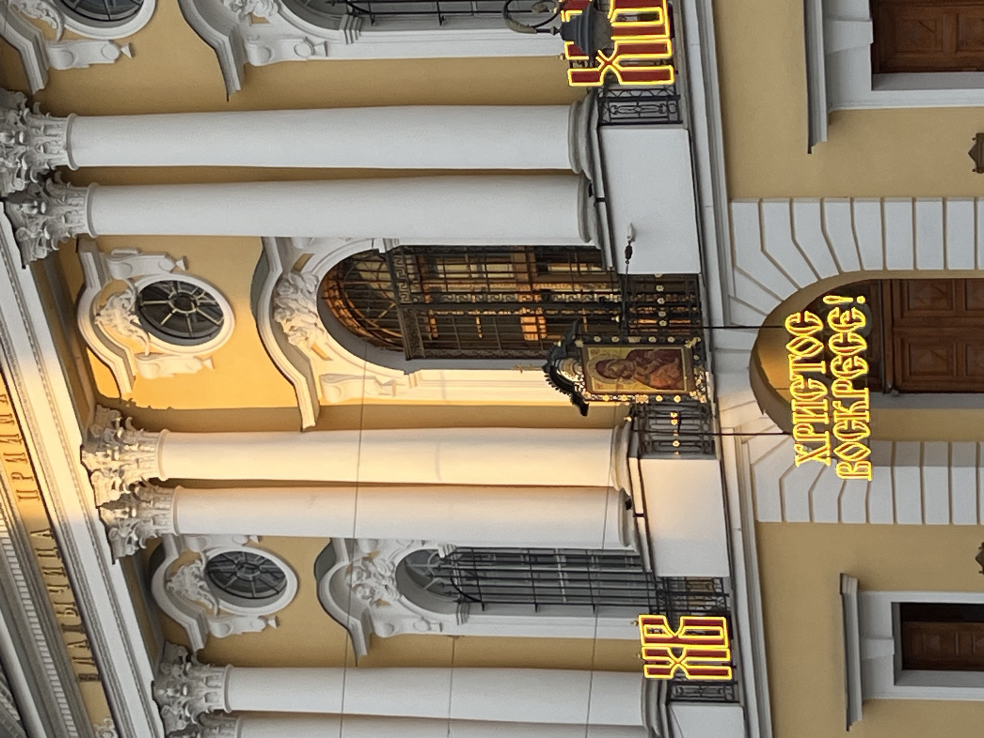 The image shows a building facade with architectural elements such as white columns, decorative moldings, and arched windows. There is a sign in Cyrillic that reads "ХРИСТОС ВОСКРЕСЕ!" in yellow neon lights, indicating the location may be Russia, Ukraine, or another country where Cyrillic is used. Above the sign is an icon. The name of the city is Unknown, and the location is Unknown.