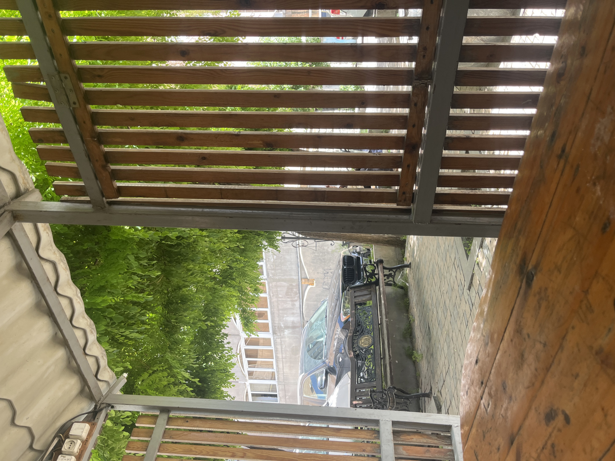 The image, taken in Yerevan, Armenia, captures a view from under a wooden pergola. The pergola is constructed of horizontal wooden slats supported by a gray metal frame. Through the slats, one can see trees, a building with windows, and a black car parked next to an ornate bench. A weathered wooden tabletop occupies the right side of the frame. Electrical outlets are visible on the lower left. The overall scene depicts a sheltered outdoor space.