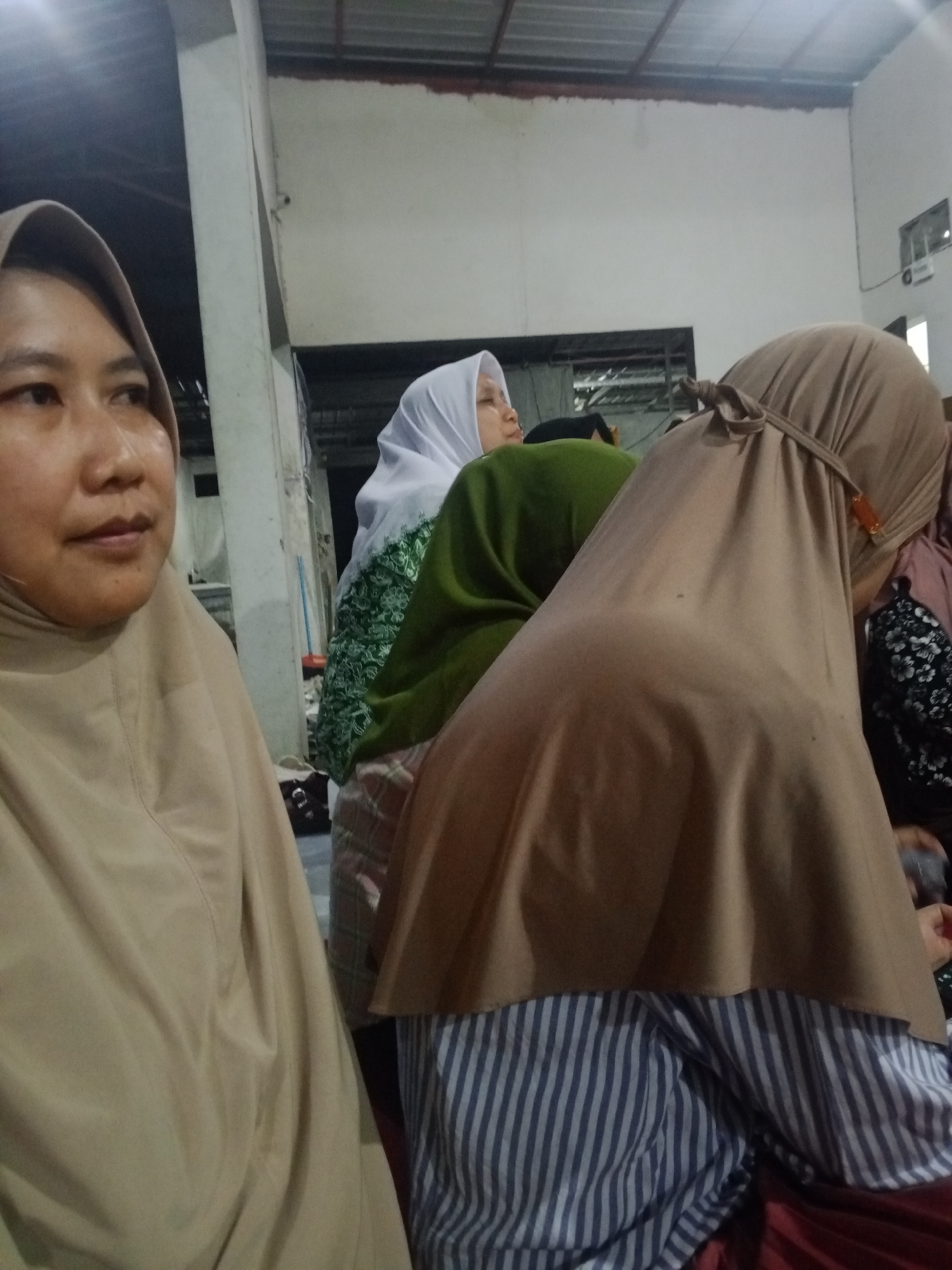 The image depicts a group of individuals indoors, likely in Mlilir, Indonesia, given the context. A woman on the left side of the frame is wearing a tan hijab and her face is partially visible, looking to the left. Behind her, other individuals are present, mostly obscured. One person is wearing a white hijab and a green patterned garment. Another person is wearing a tan hijab tied at the back with a visible orange clasp. Their lower body is covered by a striped shirt. The environment appears to be a covered space with a corrugated metal roof visible at the top. The lighting is artificial and somewhat dim, suggesting an indoor setting, possibly a gathering or event. The background shows walls and some structural elements of the building.