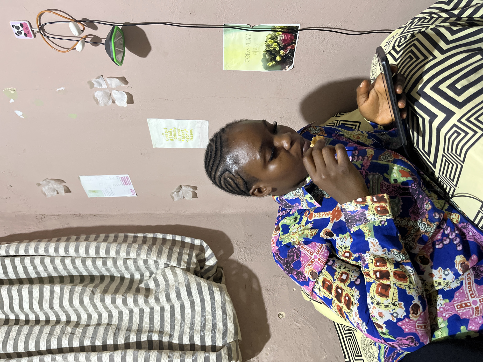 A young Black woman with her hair braided in cornrows sits on a bed with a patterned yellow and black cover. She wears a brightly colored long-sleeved shirt with a blue base and a complex pattern of pink, yellow, red, and green shapes. She is looking down, holding a piece of food to her mouth with her right hand and a smartphone in her left hand. The room has pink walls. A striped curtain hangs on the left side of the frame. On the wall to the right of the woman, there are several pieces of paper tacked up, including one with a drawing of a panda and the word "Pandy," and another with a floral image and the text "GOD'S PLAN." There is also a hanging device with headphones and what appears to be a small fan. The lighting suggests an indoor setting, possibly during the day or with artificial light. The overall scene depicts a casual moment of a person relaxing and using their phone while eating. The location is specified as Rafin Par, Nigeria.