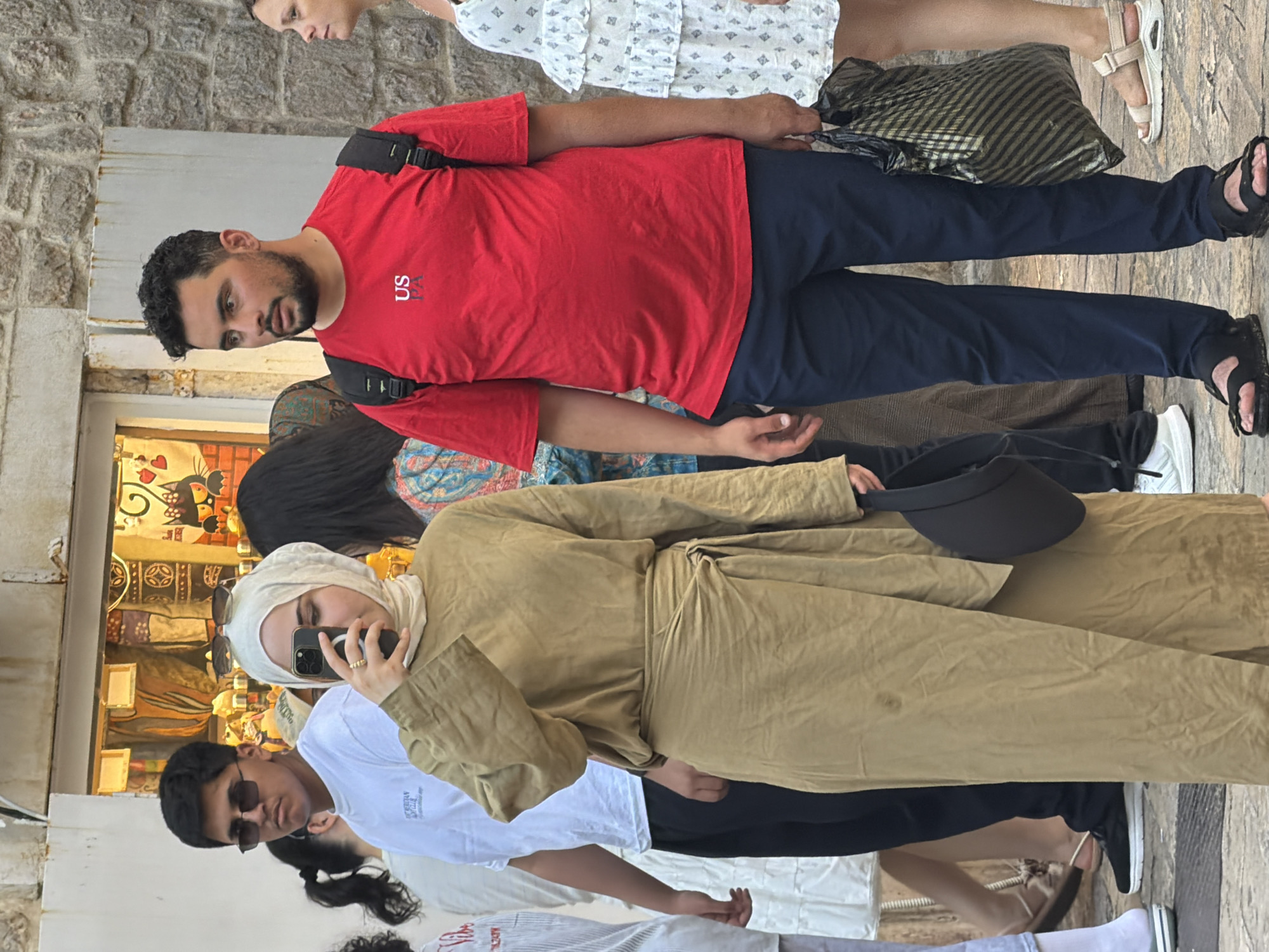In Budva, Montenegro, a street scene unfolds with several individuals. A woman in the foreground, wearing a beige hijab and a matching wrap-style dress, holds up a smartphone to take a photo. To her right, a man in a red t-shirt with "US PA" printed on it and dark blue trousers stands with a backpack. Behind them, a young man in a white t-shirt and sunglasses is visible. Further to the right, a woman in a white patterned dress walks past. The background consists of a stone building and a shop with merchandise displayed. The ground is paved, and the overall lighting suggests it is daytime.