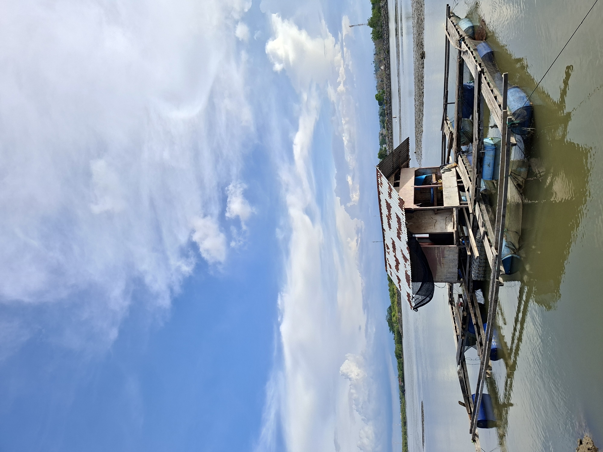 The image depicts a makeshift, two-story structure constructed on a wooden platform supported by blue plastic barrels floating on water. The structure has a corrugated metal roof with rust. The surrounding environment consists of water, likely a river or lake, with land visible in the background. The sky is bright blue with scattered white clouds. The image is rotated 90 degrees counter-clockwise. Specific details confirming this is Lhokseumawe, Indonesia, are not present in the image. Therefore, the city cannot be identified.