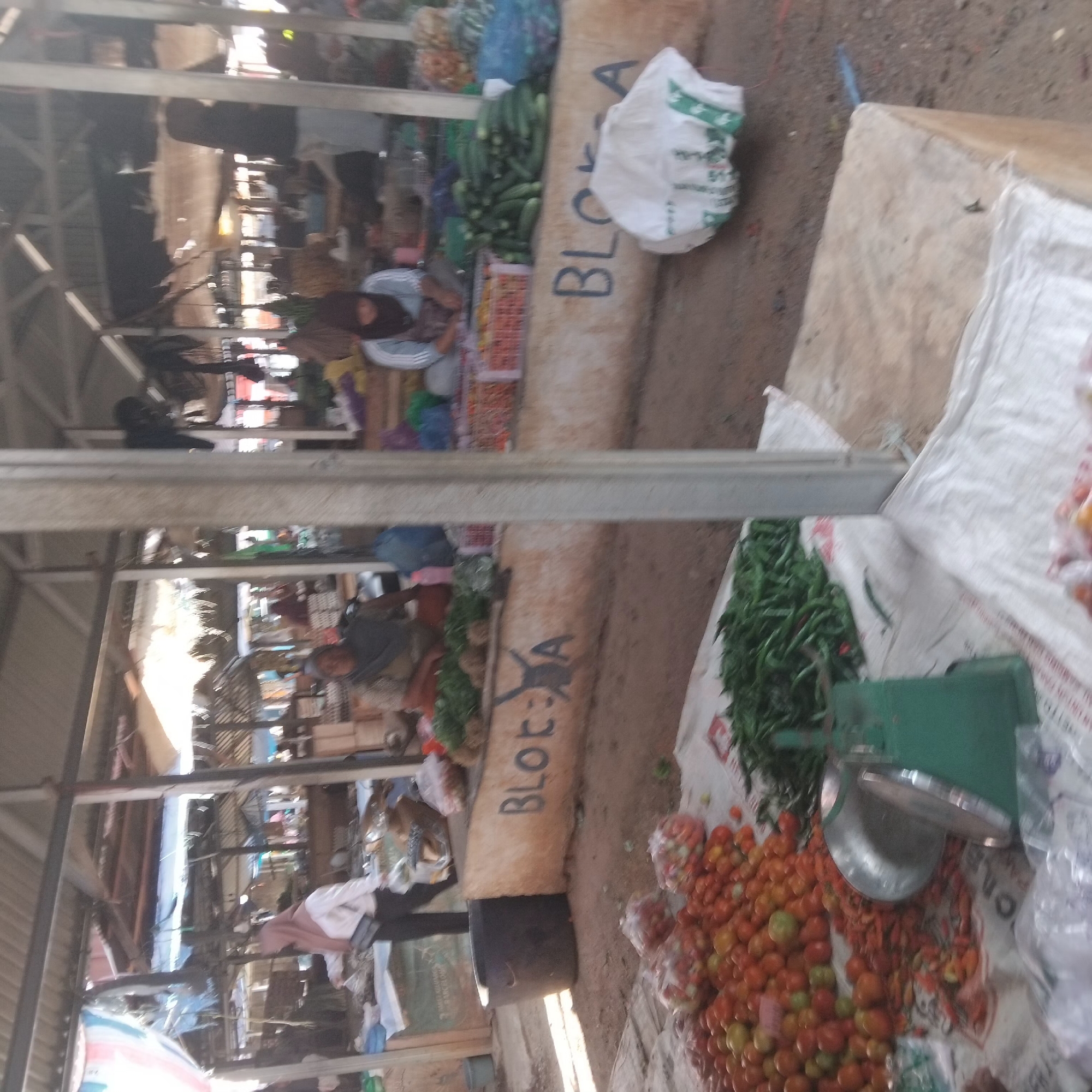 The image depicts an outdoor market scene in Pelaihari, Indonesia. A woman wearing a headscarf and a light-colored top stands near a stall laden with various vegetables. In the foreground, a pile of tomatoes is visible, alongside a green weighing scale. Several stalls with produce and other items are set up under a covered structure. The ground is dirt and sand. The overall impression is one of a busy, local market.