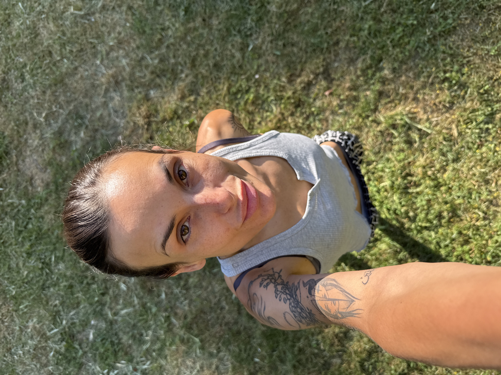 This is a first-person perspective, outdoor, daytime, high-angle selfie of a woman with brown hair pulled back and a light tan complexion, taken in a grassy area. She is wearing a light gray ribbed tank top and patterned shorts. She has tattoos on her left arm and shoulder, including a large design of branches and a bird, and a portrait. The background consists of green grass, with some dry patches and scattered sunlight. The scene does not contain any specific landmarks or indications of the city of Pleven, Bulgaria.