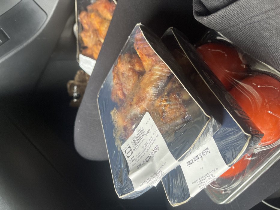 The scene is situated inside a vehicle, likely a car, in Biss'one, Switzerland. In the foreground, multiple pre-packaged food items are stacked and resting on a person's lap, which is covered by dark-colored fabric. The primary visible items include two black plastic trays of roasted chicken parts, sealed with clear plastic film. The topmost chicken tray bears a white label from "Lidl," displaying text in Italian, "alette di pollo arrosto" (roasted chicken wings), and a price of "3.99 €." Below these chicken trays, a clear plastic tray containing at least two red tomatoes is visible, also sealed with clear film. This tomato tray has a white label partially visible, which reads "costine di suino arrost..." (roasted pork ribs). In the blurred upper right background, another black plastic tray containing roasted chicken parts is partially discernible. The vehicle's interior appears dark, with indistinct dark grey or black upholstery and vehicle components visible. No faces or specific human actions are depicted.