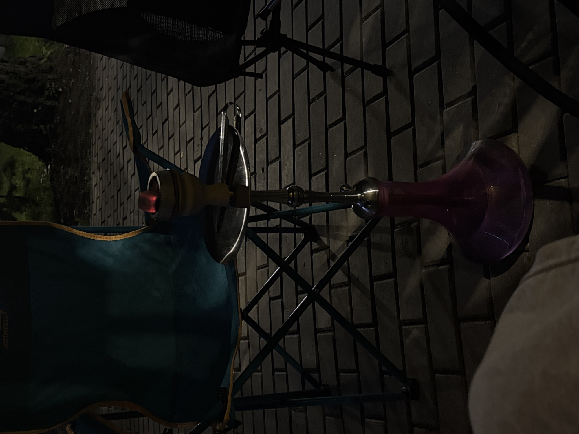 An outdoor scene at night shows a hookah setup on a brick-paved surface. A teal folding chair is positioned to the left, with a dark object, possibly another chair, to the right. The hookah is made of metal with a glass base filled with purple liquid. A red glowing coal rests on top of the hookah bowl, indicating it is in use. The ground is paved with rectangular bricks. A large tree is visible in the background, with foliage and darkness suggesting it is nighttime. The location is specified as Doma 1096 Km, Russia.