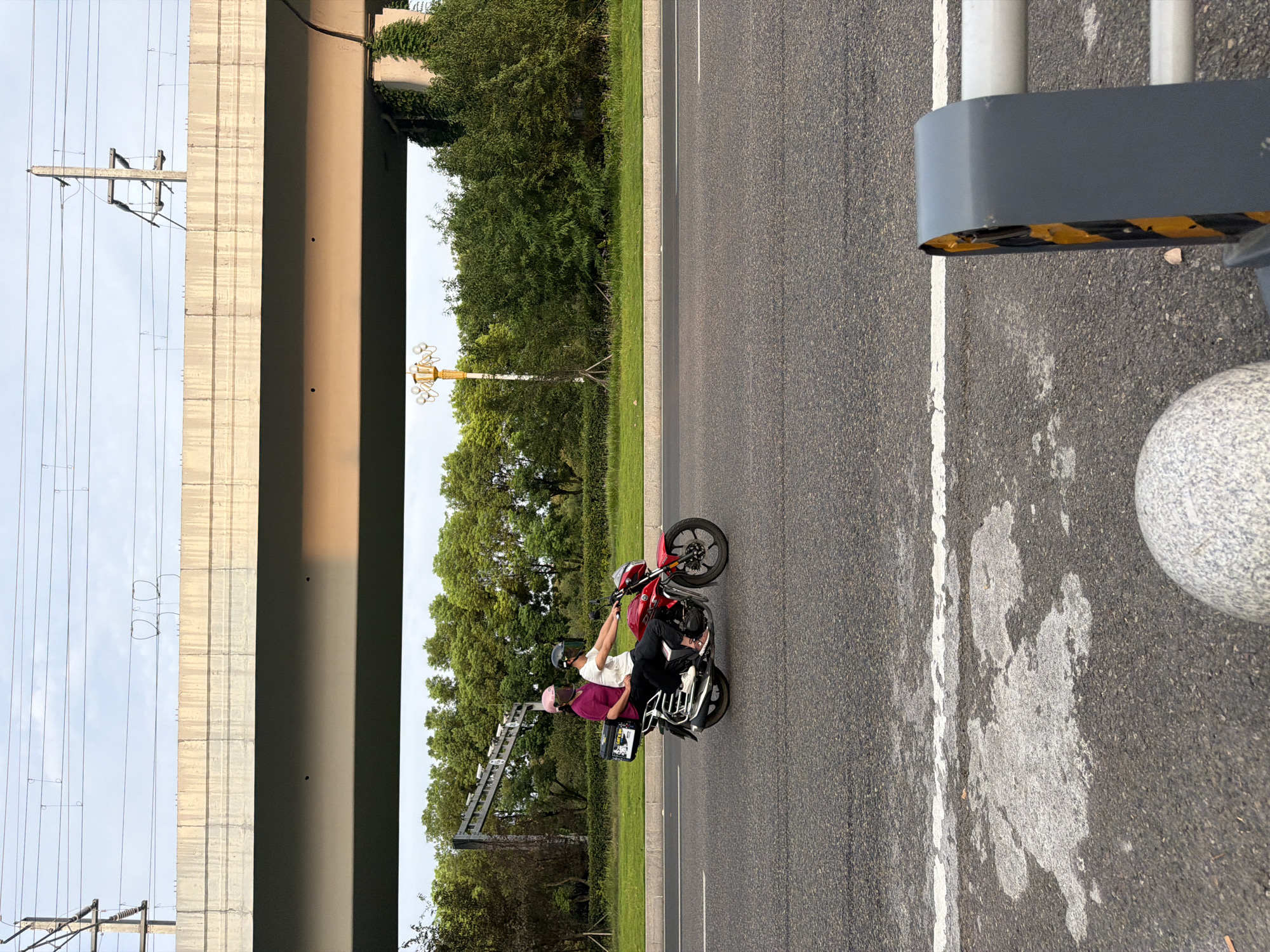 Two people are riding a red motorcycle on a paved road under an elevated highway. The rider in front is wearing a white shirt and a black helmet with a visor, and the passenger behind is wearing a purple shirt and a pink helmet. The motorcycle has a black storage box attached to the back. The road is marked with white lines, and there are some white splotches on the asphalt. The background consists of green trees, grass, and a concrete barrier. Overhead, power lines and the underside of the highway structure are visible against a hazy sky. The scene is set in Jiangjin, China, as indicated by the surrounding environment typical of urban infrastructure in that region.