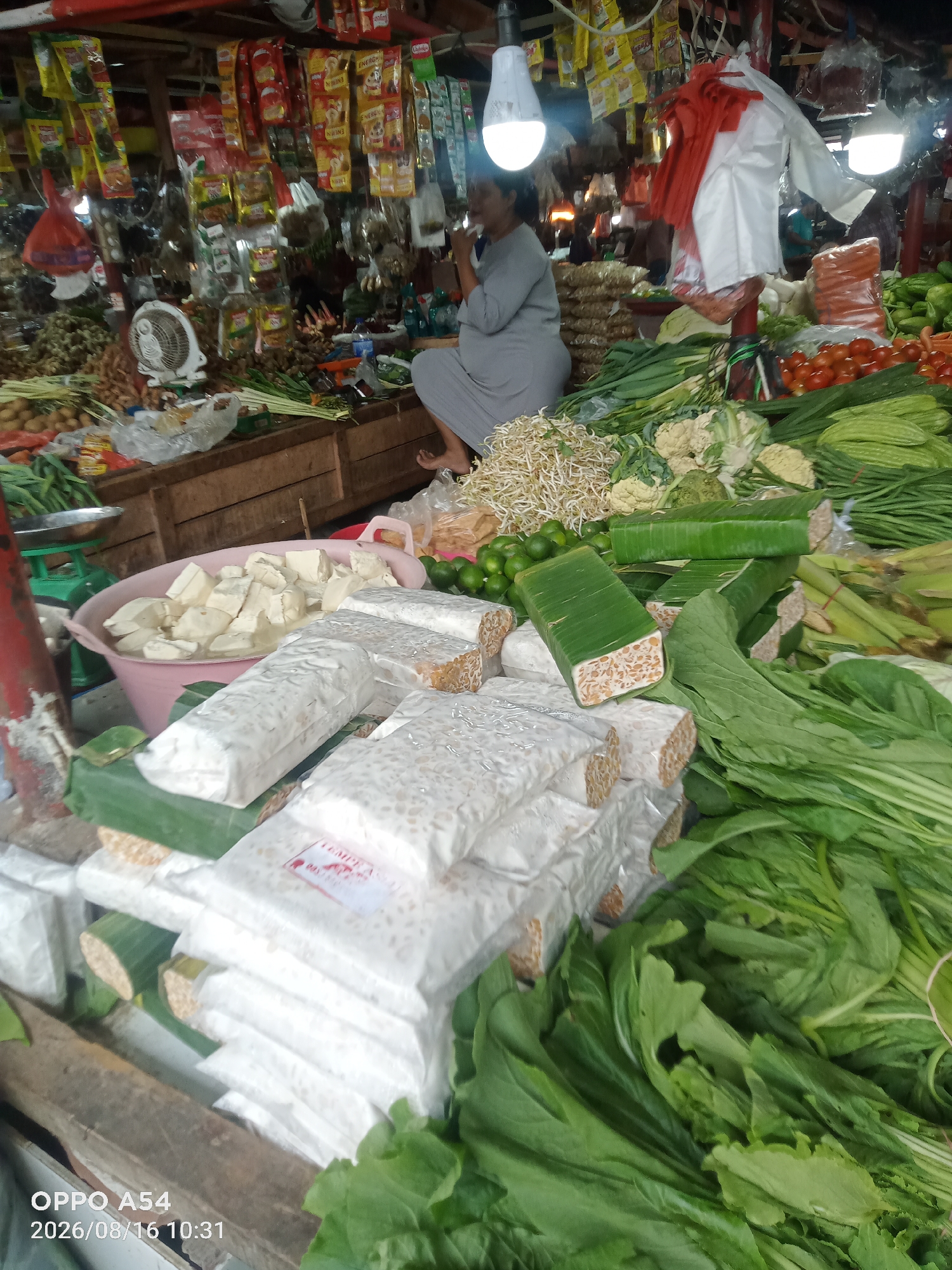 The image depicts a bustling outdoor market scene, likely in Jambi, Indonesia, given the timestamp on the lower left. A woman in a grey outfit is seated behind a stall laden with fresh produce and packaged goods. In the foreground, several large packages of tempeh, a fermented soybean product, are prominently displayed, some wrapped in plastic and others in banana leaves. Adjacent to the tempeh are tubs of cubed tofu submerged in liquid, and further back, a variety of vegetables including bean sprouts, limes, cauliflower, chilies, and leafy greens are visible. The stall itself is constructed from wood, and overhead, numerous packaged food items and a fan hang from the market's canopy. The lighting suggests it is daytime, with artificial lights also illuminating the stall area. The overall impression is one of a vibrant, local marketplace with a diverse array of fresh food offerings.