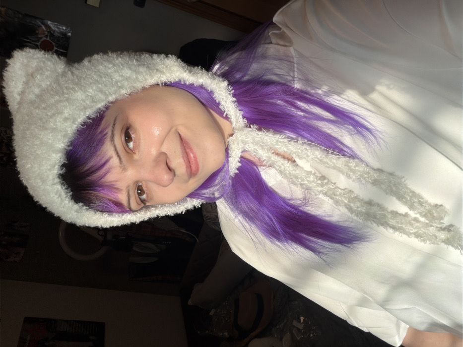 The scene depicts a single individual, appearing female, in an indoor setting. The specific location within Sof'ino, Russia, is not discernible from the image content. The individual is positioned centrally, facing slightly right of the camera, and looking directly at the viewer with a subtle smile. She has long, straight, vibrant purple hair with bangs, light skin, and brown eyes. She is wearing a fluffy, light-colored (appearing white or off-white) hat with two rounded, ear-like protrusions on top and two matching fluffy ties hanging down the front. Below the hat, a light-colored (white) collared shirt or blouse is visible. Strong, natural light, likely sunlight, illuminates the subject from the left, creating highlights on her skin and clothing, and casting the background into partial shadow. The background, largely out of focus, reveals a dark wall, a partial view of a white circular object (possibly a ring light), indistinct posters or artwork, a dark sofa or bed with light-colored pillows, a woven basket-like object with dark stripes, and some items in plastic packaging.