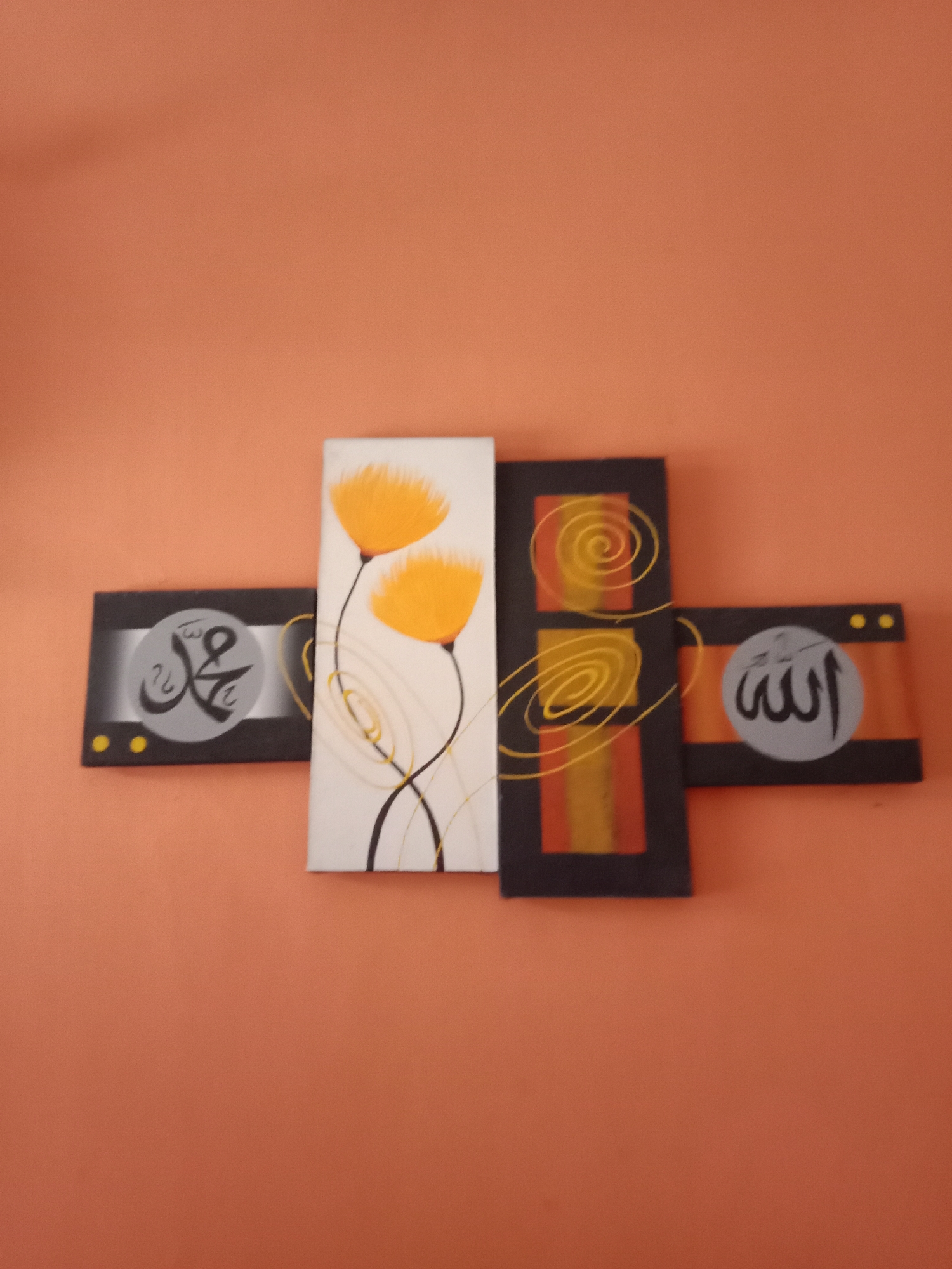 This is a photograph of a four-panel Islamic calligraphy artwork displayed on an orange-toned wall. The artwork is horizontally arranged and consists of two narrow side panels and two wider central panels. The left narrow panel features Arabic calligraphy within a grey circular emblem, set against a black background with subtle gradient effects. Two yellow dots are placed below the emblem. The first central panel is a vertical rectangle with a white background, depicting two abstract yellow flowers with black stems and decorative yellow swirls. The second central panel is a vertical rectangle with a dark background, featuring vertical stripes of red and yellow, and two prominent yellow spiral designs. The right narrow panel has an orange and black striped background with two yellow dots above a grey circular emblem containing Arabic calligraphy. The overall impression is of decorative religious art. The location is specified as Kutosari, Indonesia.