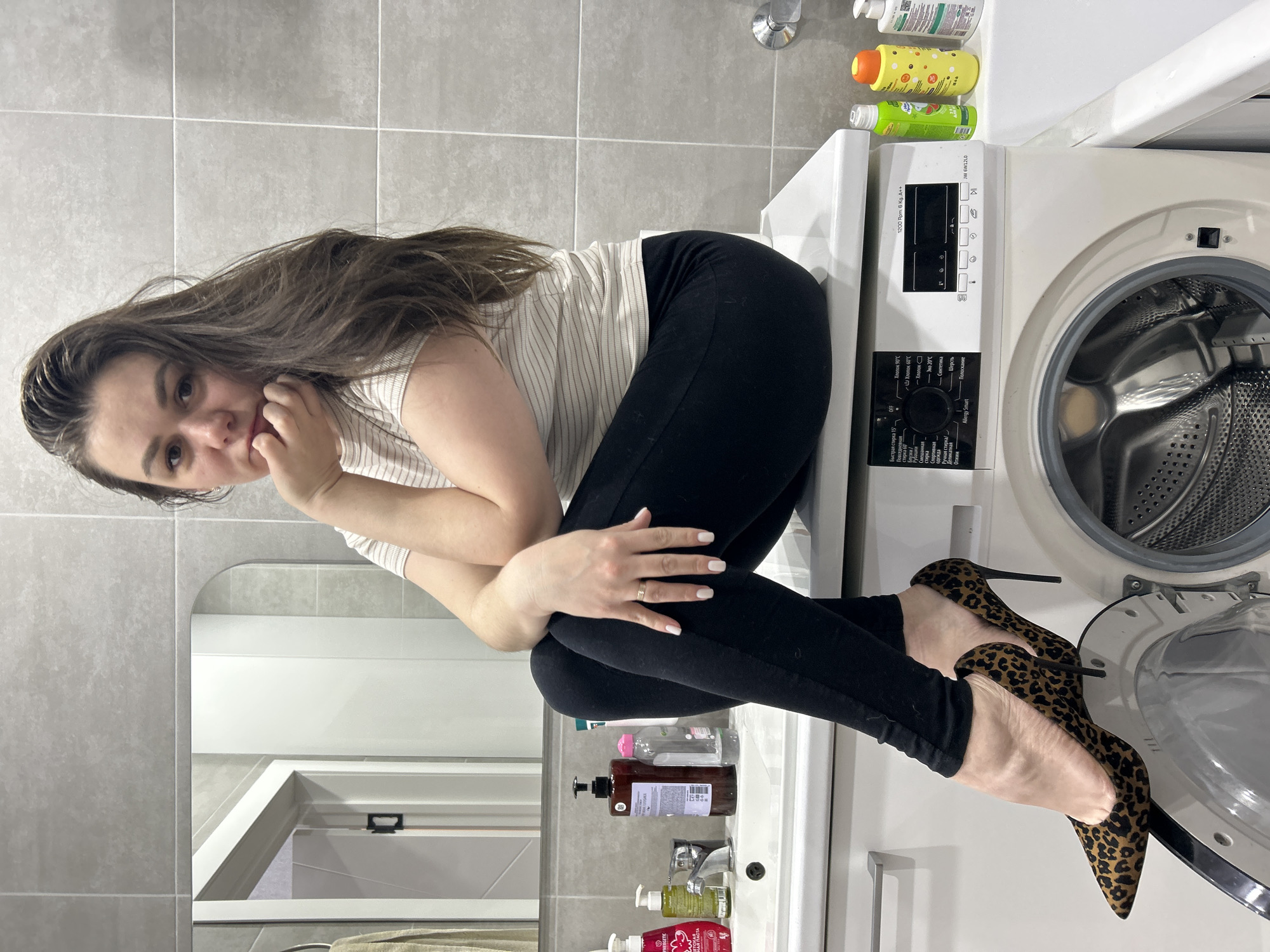 The image displays a young woman sitting on top of a white front-loading washing machine in a modern bathroom, located in Chaikovsky, Russia. She has long, dark brown hair and is wearing a light-colored striped t-shirt, black pants, and striking leopard print high heels. Her pose is contemplative, with her right hand resting near her chin, and her gaze directed towards the viewer. Her left arm is crossed, and she wears a simple ring on her left hand, with white nail polish visible.
The washing machine's door is slightly ajar, revealing its metallic drum. Its control panel features a central knob and several buttons, along with a digital display. On the counter to the left of the washing machine, above a sink, various bottles of soap and shampoo are arranged. A mirror hangs above the sink, reflecting parts of the room. The bathroom walls are covered in light grey square tiles, giving the space a clean, minimalist feel.
Visible text on the washing machine includes "1200 Rpm. 6 Kg. A++" and the model number "JW 6W12L0". Numerous program options are listed in Cyrillic, such as "Быстрая стирка 15'" (Quick wash 15'), "Хлопок 90°" (Cotton 90°), "Синтетика" (Synthetics), "Шерсть" (Wool), "Полоскание" (Rinse), "Отжим" (Spin), and "Allergy Smart". Other visible program labels include "Хлопок 60°С", "Эко 20°С", "Рубашки" (Shirts), "Смешанная стирка" (Mixed wash), "Спортивная одежда" (Sportswear), and "Ручная стирка" (Hand wash). The word "YNERGETIC" is also partially visible on a bottle. The scene captures a moment of casual posing with a touch of personal style, set against the backdrop of an everyday domestic environment.