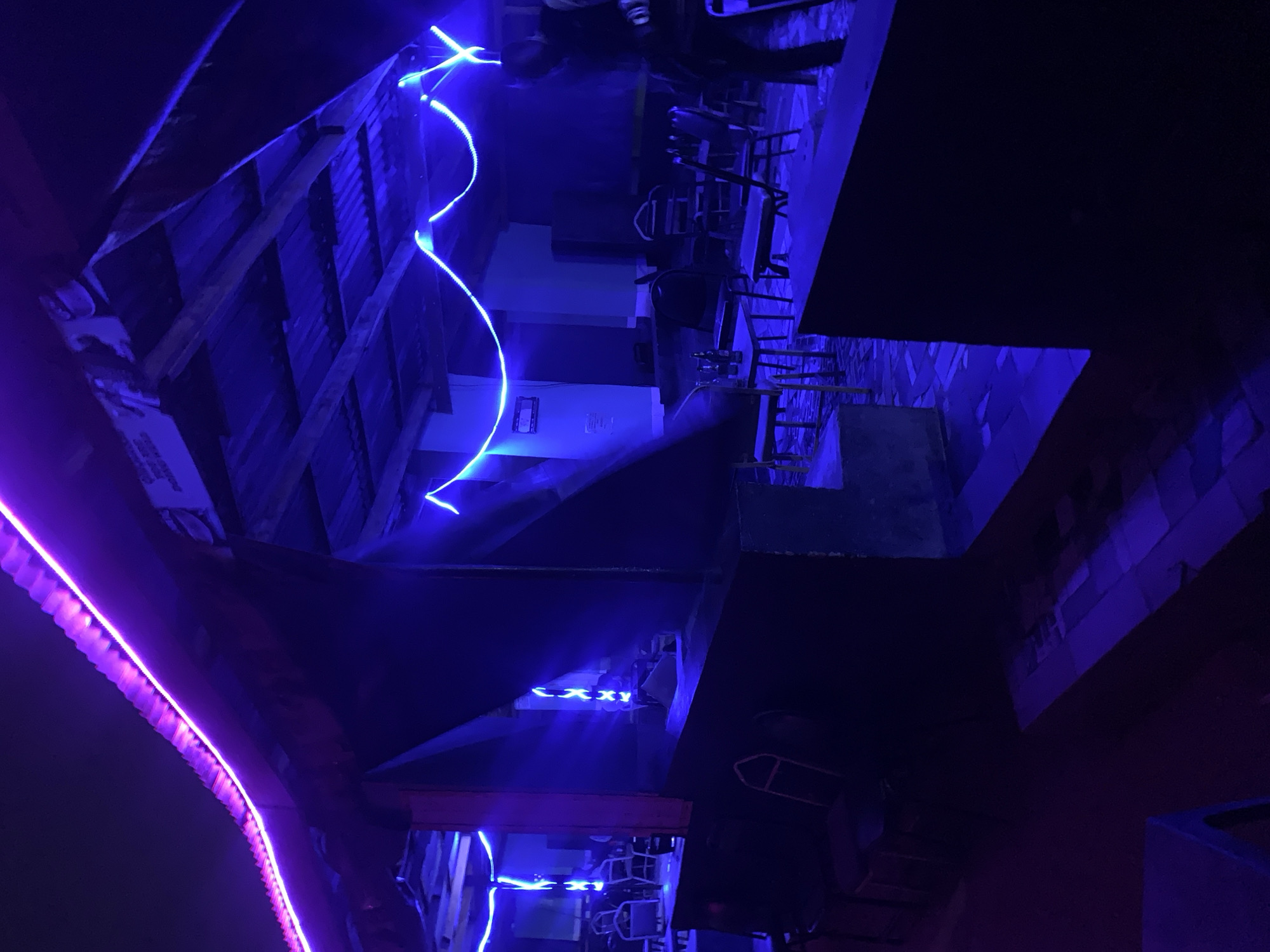 The image shows an interior scene with blue and purple neon lighting. There appear to be tables and chairs, suggesting a restaurant or bar. The architecture consists of corrugated metal and wooden beams, with the neon lights highlighting these features. Some individuals are faintly visible in the background, though their actions are obscured by the darkness and limited detail. The setting is likely in New Stadium, Nigeria.