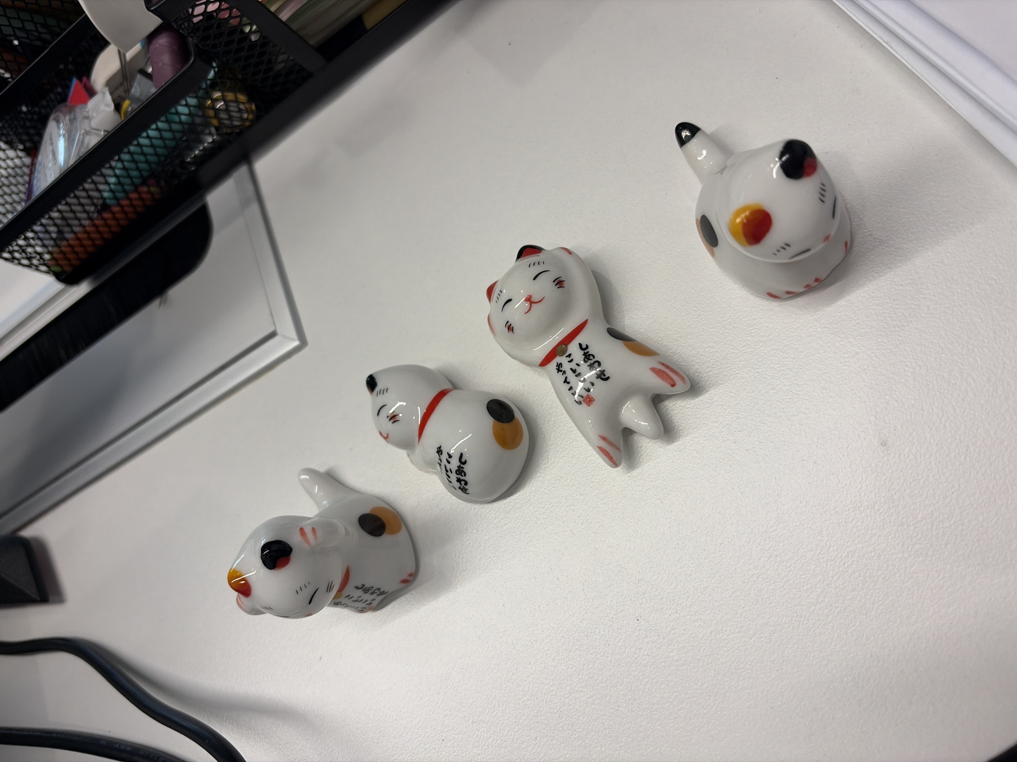The image shows a top-down view of a white desk surface. On the right side of the desk are four ceramic figurines: one is a rounded cat head with a black and orange spot and a tail, another figurine shows a cat sitting up. The two last figurines are laying down on their belly. Each one has white ceramic base with red accents. To the left of the desk is a black wire mesh container. Underneath the desk, a black power cable is visible. No evidence suggests the location of this scene.