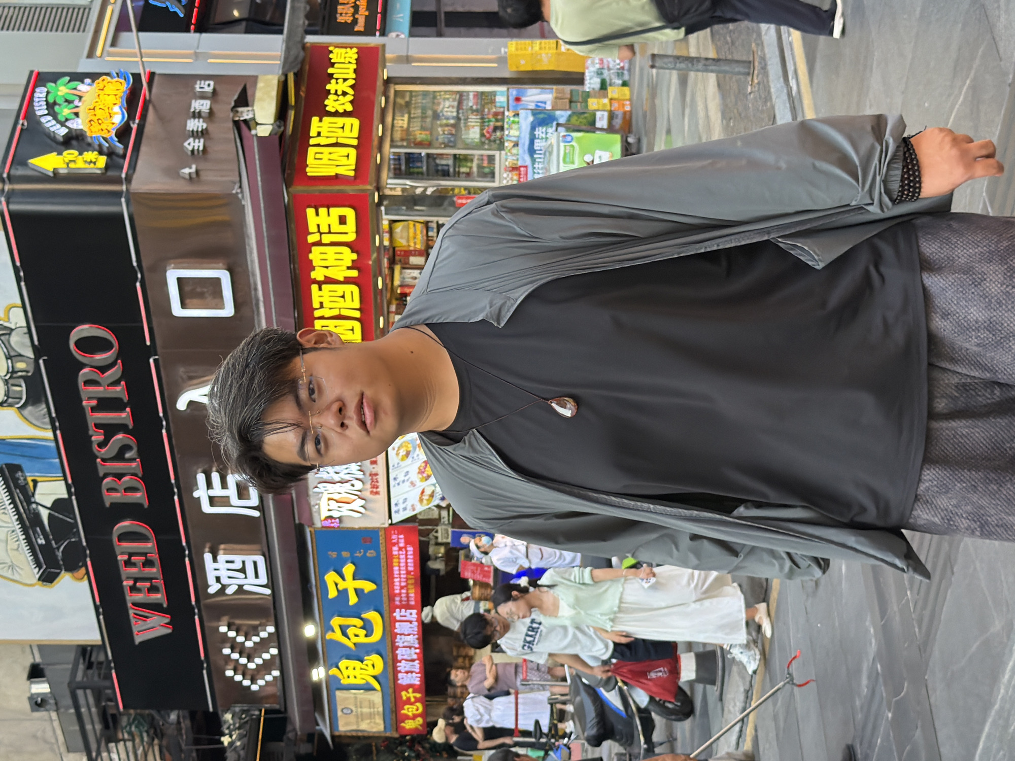 In Yuzhong District, China, a young East Asian male stands center-frame in a street scene. He is wearing glasses, a dark t-shirt with a pendant necklace, a grey unzipped jacket, and patterned pants. His right hand, adorned with a beaded bracelet, rests at his side. Behind him, a bustling street is visible with various shops and pedestrians. A prominent sign reads "WEED BISTRO" in red lettering. Other signs in Chinese characters are also present, including those for "酒 店" (hotel) and "鬼 包子" (ghost buns). In the background, people are walking, and a motorcycle is parked to the left. The environment appears to be an urban commercial area with a lively atmosphere.