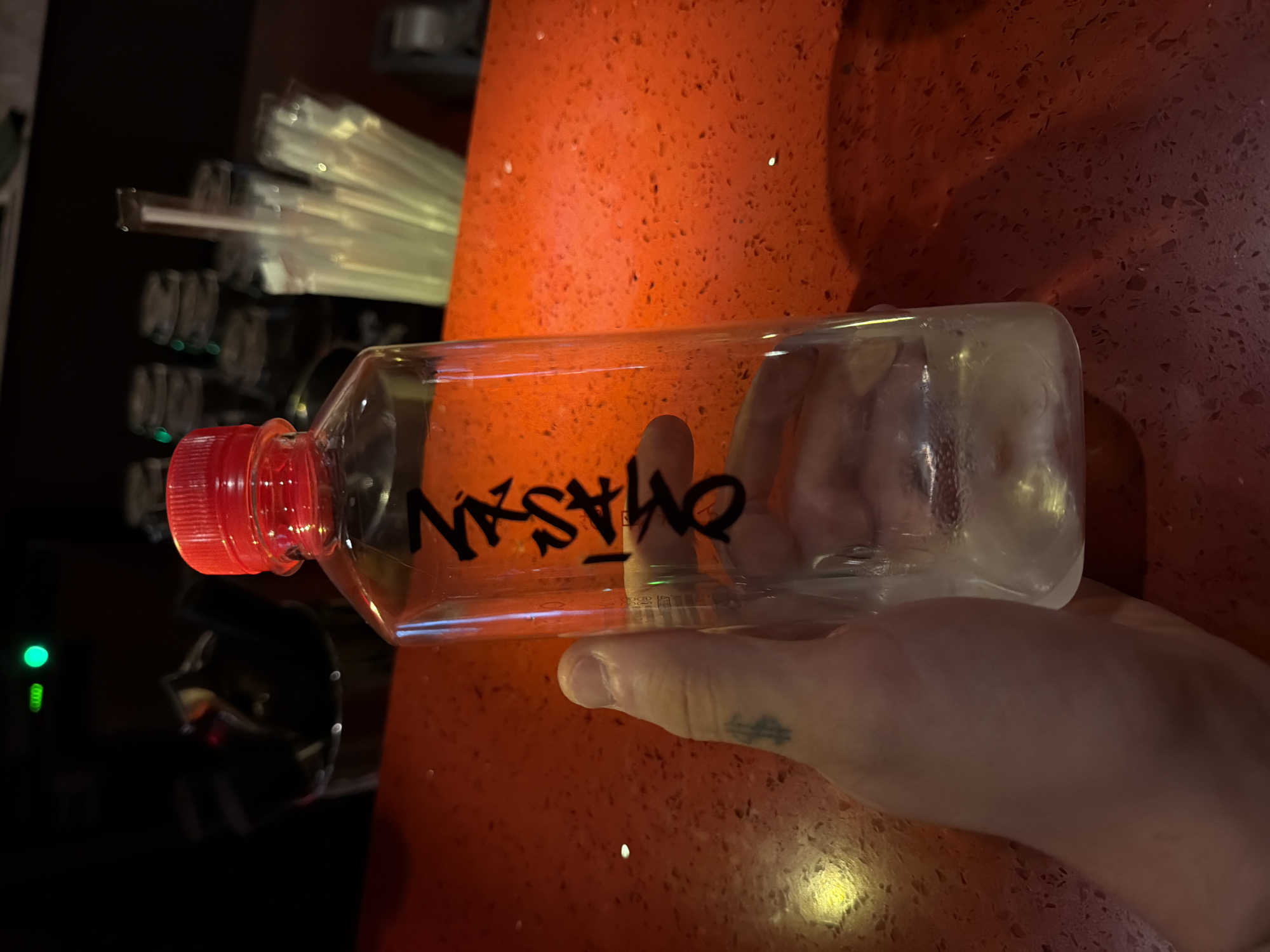 A clear plastic bottle with a red cap, filled with a clear liquid and ice, is held in a person's left hand, which has a dollar sign tattoo on the thumb. The bottle has black writing on it that appears to be Japanese characters. The bottle is placed on a bright orange, textured countertop, likely in a bar or restaurant setting. In the background, out of focus, are a metal container with utensils and a stack of plastic straws. The lighting suggests an indoor environment, possibly at night, with a green light visible in the upper left corner of the frame. The scene is static, with no active interactions depicted. The location is not specified as Bangkok, Thailand.