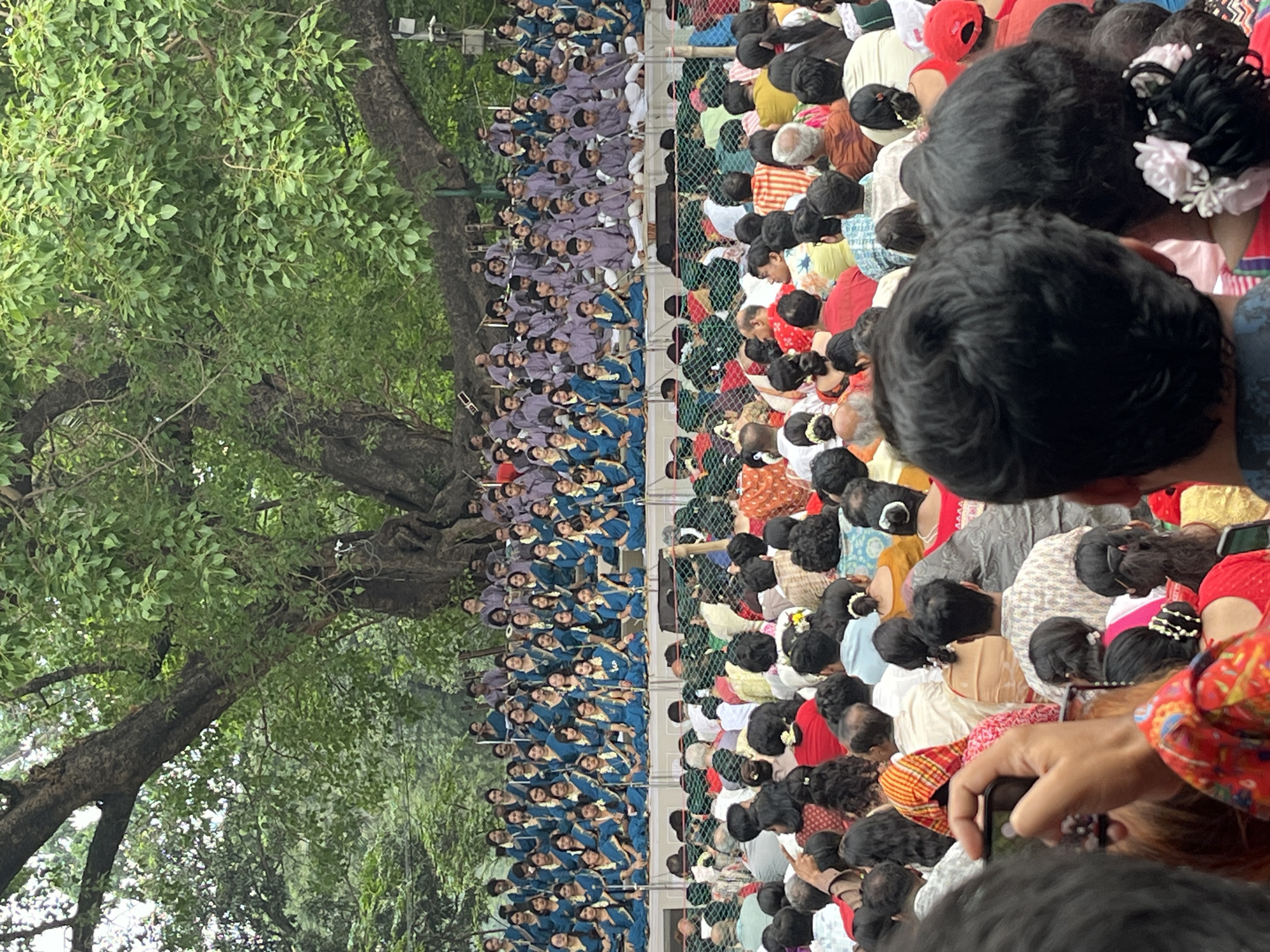 The image shows a large gathering of people, likely in Dhaka, Bangladesh, given the cultural attire and appearance of the individuals. On the left, a large group of people wearing similar blue and grey clothing are sitting in rows. To the right of a fence, a large crowd is sitting, appearing to be observing the people in blue. In the foreground, a person is taking a photograph. A large tree is visible to the left.