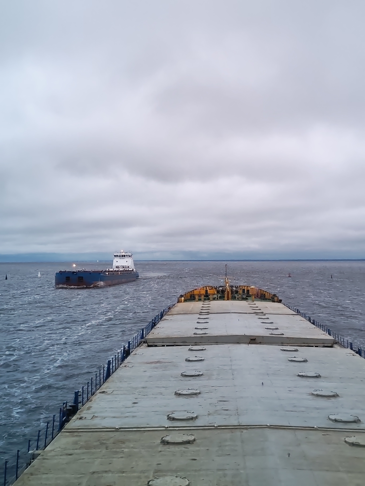 The image depicts a view from the deck of a large ship, looking out towards another ship in the distance. The sky is overcast with gray clouds. The water is choppy with small waves. The ship in the foreground has a light gray deck with rectangular panels and oval hatches. The second ship is blue, with a white superstructure. There are buoys in the water, one on each side of the ships. The location cannot be identified as Bereg, Russia, based on the visual information provided.