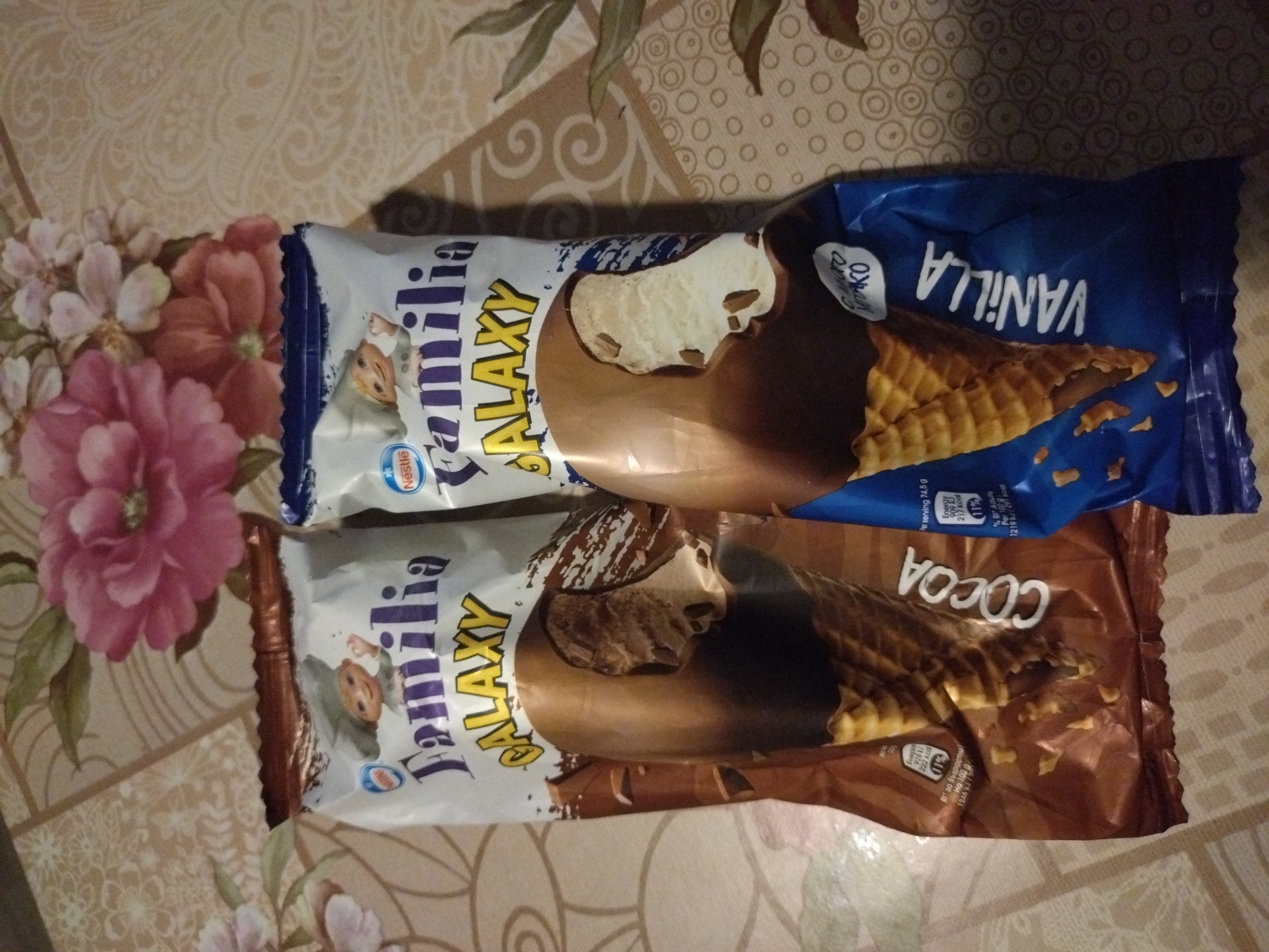 The image shows two Nestle Familia Galaxy ice cream bars. One is vanilla flavored and packaged in a blue wrapper, the other is cocoa flavored and packaged in a brown wrapper. Both are lying on a table with a floral patterned tablecloth. There is no information in the image to indicate the location is Stara Zagora, Bulgaria.