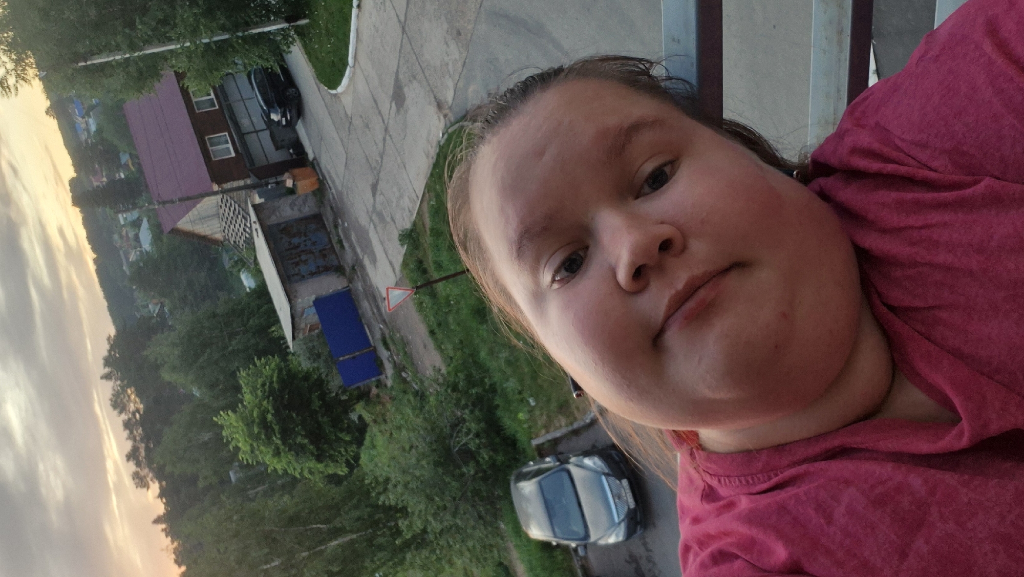 A young woman is taking a selfie. She is wearing a pink shirt. The background shows a street with houses, trees, and cars. There is a road sign visible. It could be the city of Chaikovsky, Russia.