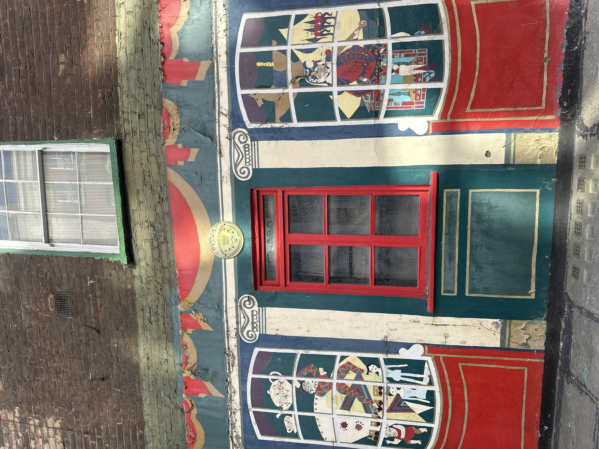 The image shows a mural on the side of a building in London, United Kingdom. The mural depicts theatrical scenes framed by painted windows and red theatrical curtains. In the center is a red-framed window. To the left and right of the central window are two large, multi-paned windows, each filled with colorful depictions of characters and objects. The left window shows figures in what appears to be a domestic or tea-time setting, with a teapot and teacups visible. The right window features more theatrical figures, including a jester-like character and a procession of soldiers. The mural is painted on a teal green background, with red and gold decorative elements. Above the mural, a brick wall is visible, with a small, white-framed window and a green trim. The mural itself shows signs of age and wear, with peeling paint. Below the mural, a red base is painted, with gold trim, and below that, a dark green section with gold trim. The scene is lit by sunlight, creating shadows on the wall. The street in front of the building is paved with cobblestones and has a metal grate.