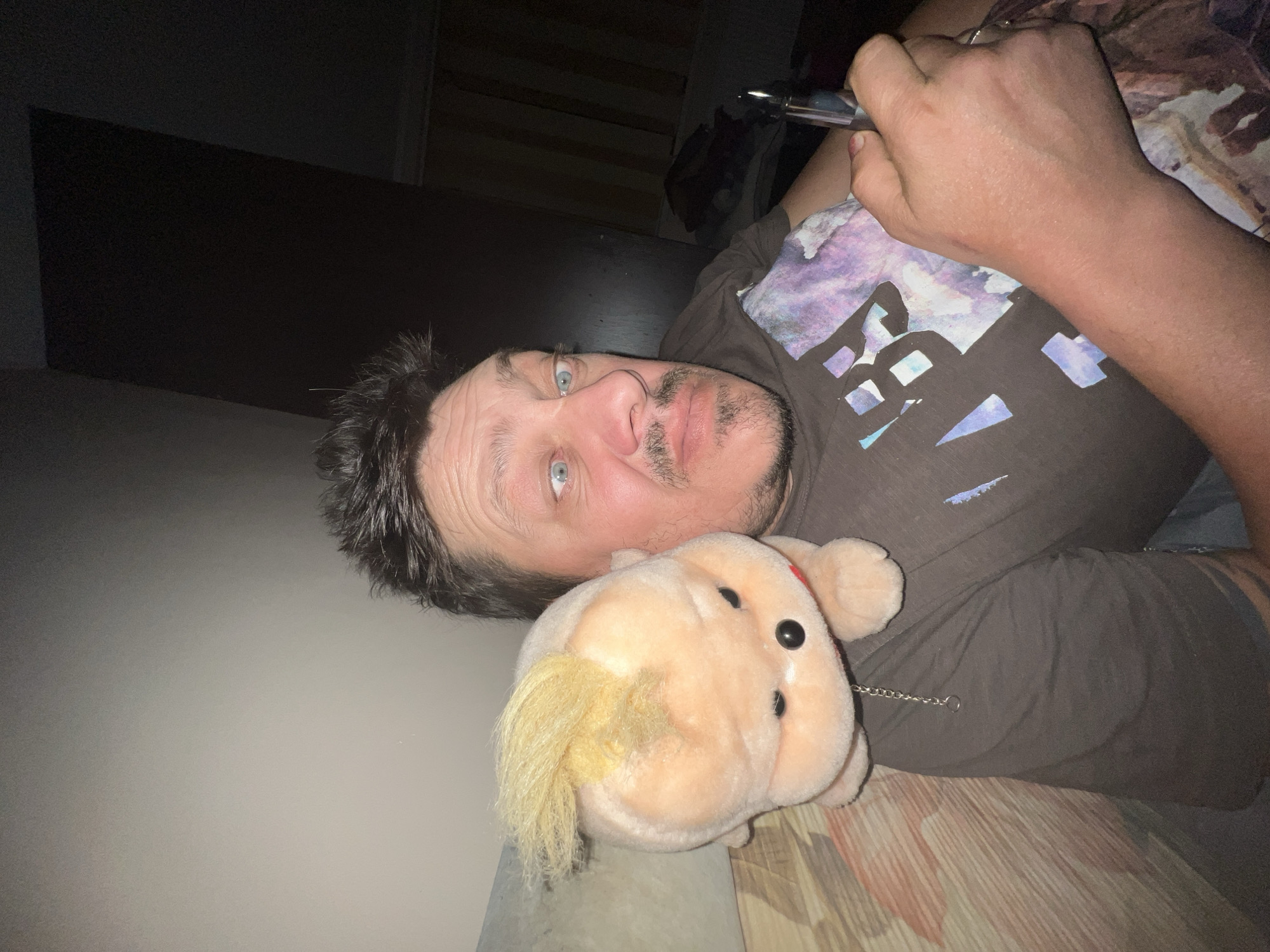 In an indoor dimly lit environment, a man is shown with a stuffed toy resting on his shoulder. The man's face is turned towards the camera, with his eyes wide open and looking slightly to his right. He has dark, tousled hair and a light mustache and goatee. He is wearing a dark t-shirt with a colorful, abstract design on the chest. In his right hand, he is holding a silver colored object, possibly a pen or vape. The stuffed toy appears to be a light-skinned creature with blond hair styled into a mohawk and black button eyes. A silver chain is attached to the toy, hanging down its front. The background shows a wall to the left and a darker area to the right, with some draped fabric visible. The scene is likely captured in a residential setting. The provided context indicates the location is the city of Drohobych, Ukraine.