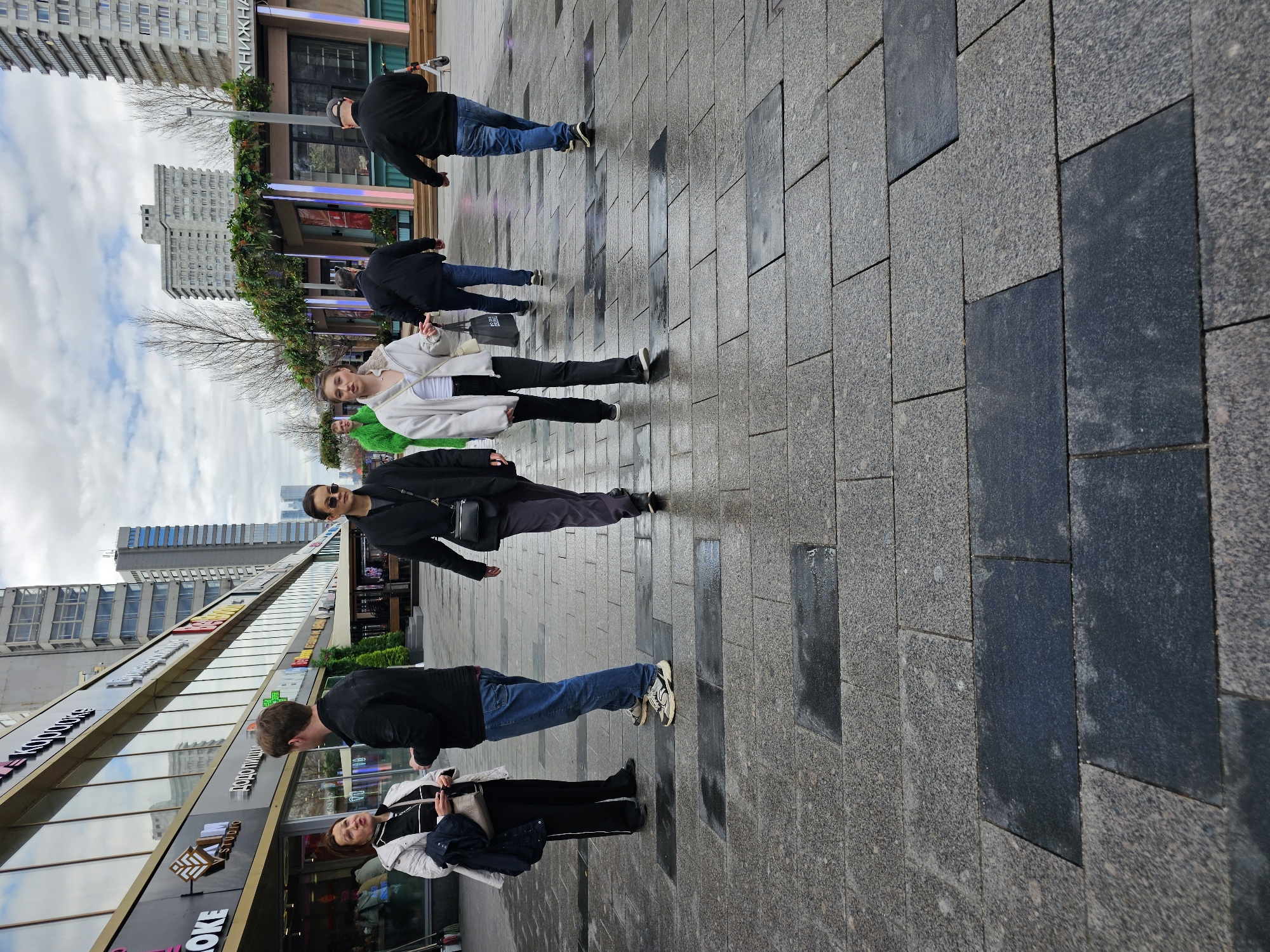 The image shows a street scene in what appears to be Moscow, Russia, based on the architectural style of the buildings. The view is along a pedestrianized area with a paved surface of grey and dark grey rectangular stones. There are six people visible in the shot, all appearing to be walking.
On the left side of the frame, there's a multi-story building with large glass windows displaying various shops or businesses.
In the background are additional buildings, including high-rise apartment complexes.
The sky is overcast with a mix of grey clouds. The scene suggests an urban environment with commercial activity.
