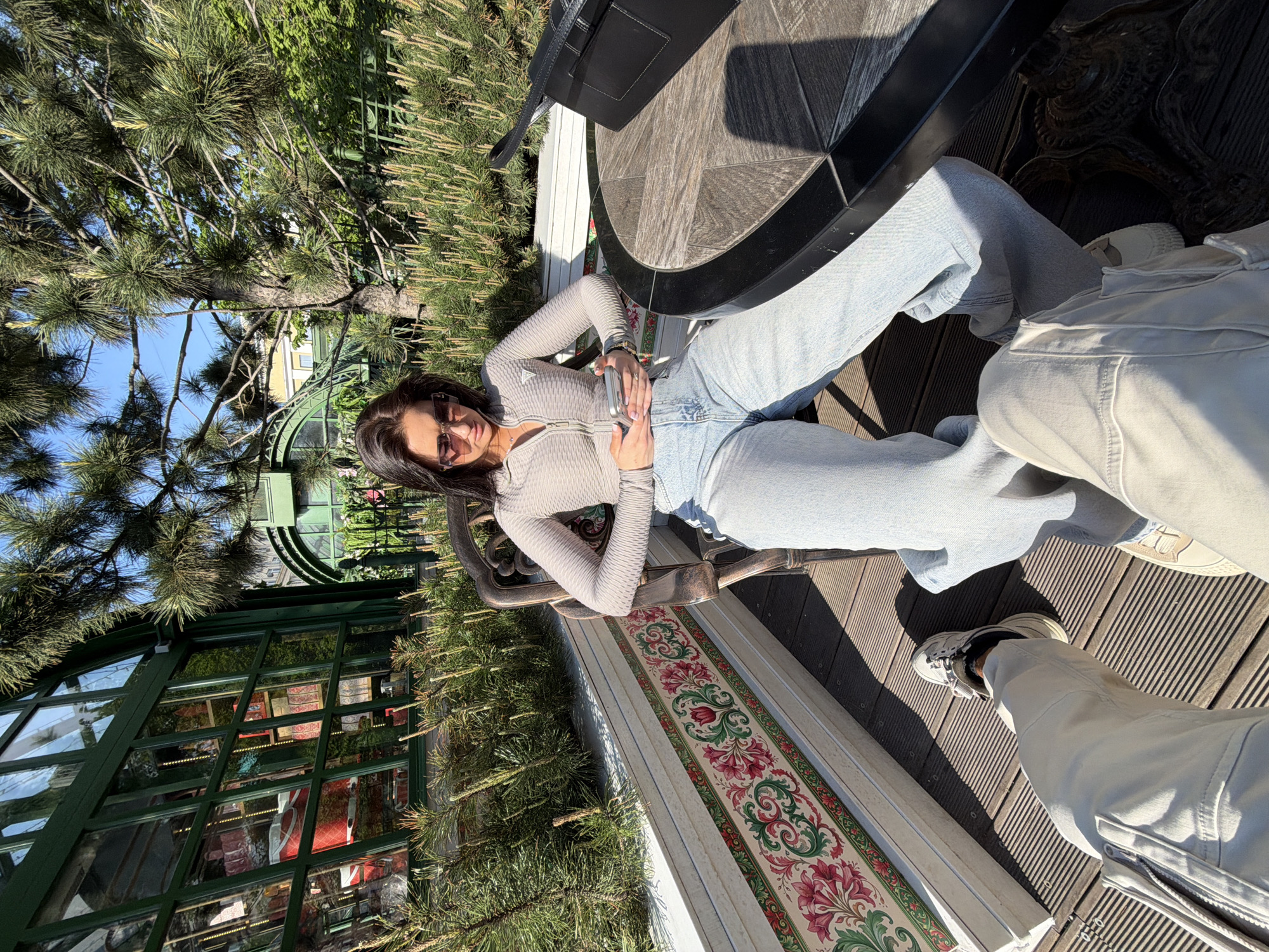 The image captures a scene set outdoors, likely in a garden or park setting. A woman with dark hair and sunglasses is positioned in the center, seated on a bench with a colorful floral pattern. She wears a gray ribbed top and light blue jeans. Next to her is a round table, likely made of wood with a black metal base. A black bag is placed on the table.
To the right of the woman, a person's legs clad in light gray pants and sneakers are visible. In the background, there's a green glass structure, potentially a greenhouse or conservatory, partially obscured by trees. The scene is bathed in sunlight, creating distinct shadows on the wooden deck. The location might be in Moscow, Russia, based on the architectural style of the glass structure. The composition suggests a candid or posed moment in a relaxed outdoor setting.