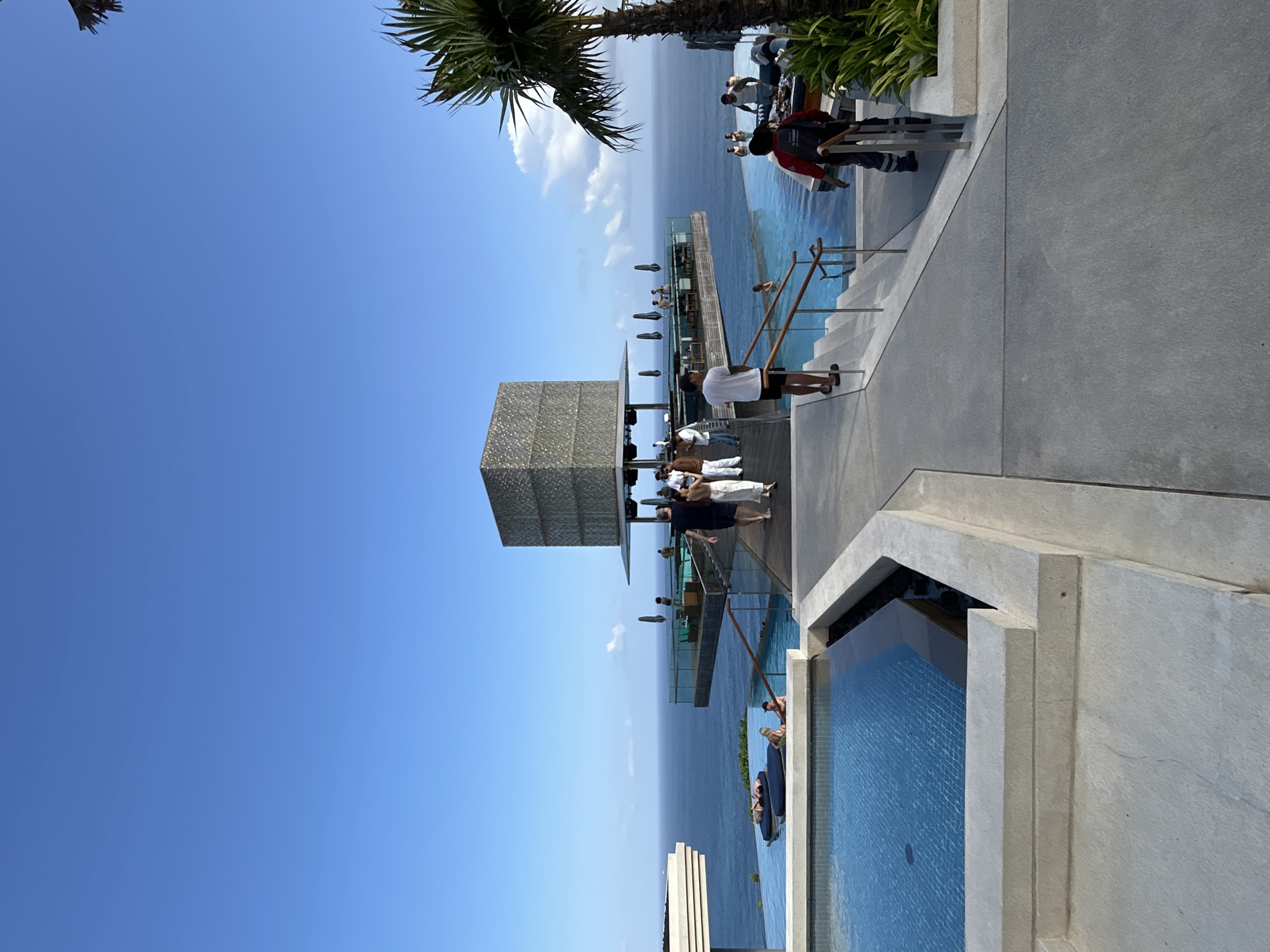 The image shows a modern resort with an infinity pool overlooking the ocean. The resort appears to be located on a cliffside. In the foreground, there is a large concrete area with a swimming pool to the left. Further back, there is a walkway leading to a multi-story structure with a cube-shaped building on top. Several people are seen walking on the walkway and in the pool area. The ocean is visible in the background, with a clear blue sky above. The architectural style is contemporary, with clean lines and geometric shapes. The location is identified as Kuta Selatan, Indonesia.