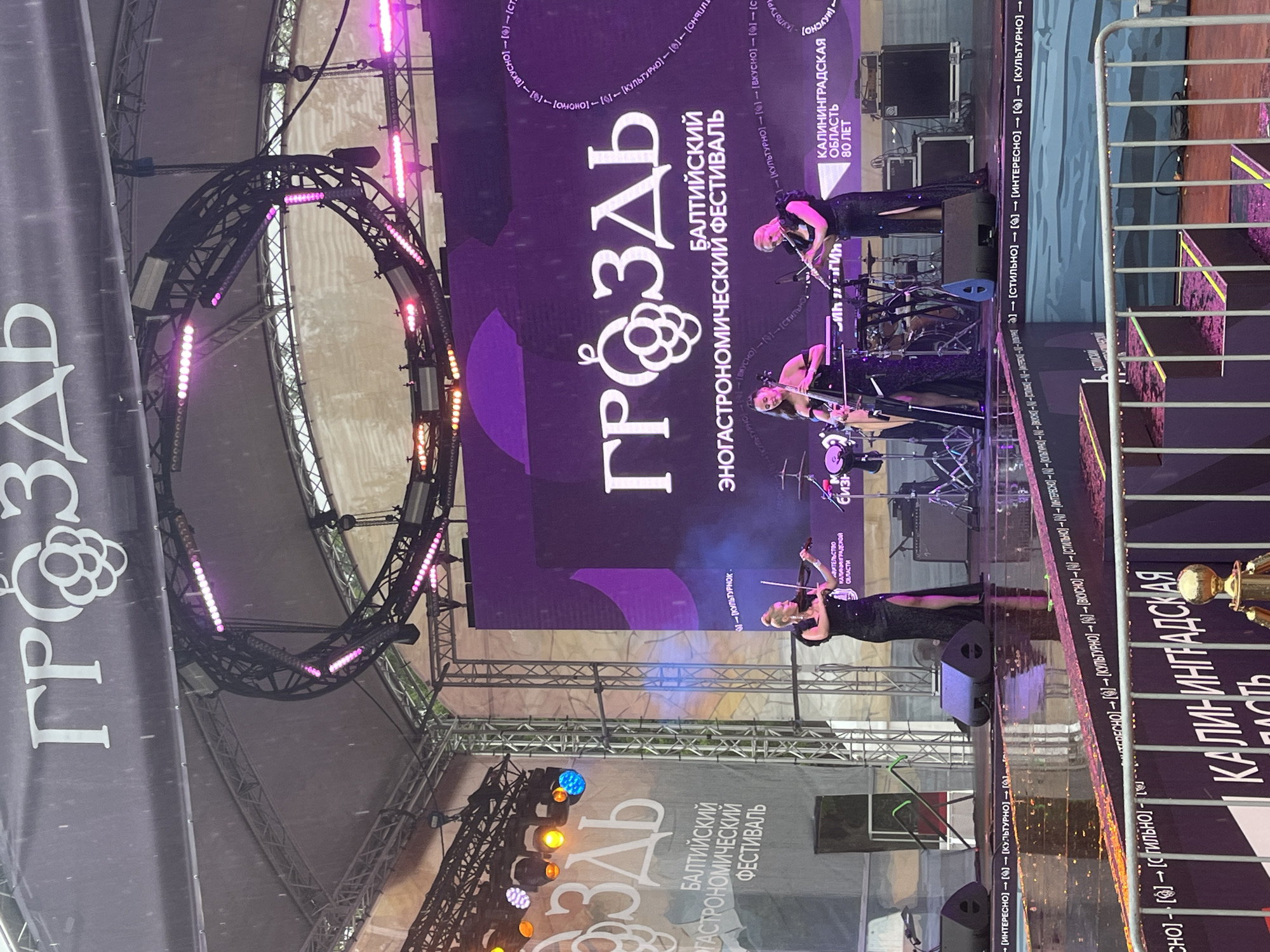 The image captures an outdoor stage performance in Svetlogorsk, Russia, likely during the "Grozdy" (Grapes) Gastronomic Festival. Three women are performing on a raised stage equipped with a large purple LED screen displaying the festival's logo and name. The woman on the left is playing a violin, dressed in a long black sparkly dress with a slit. The central performer, also in a similar dress, is playing a flute. The performer on the right is playing a cello. The stage is wet, indicating it is raining, with reflections visible on the floor. Multiple speakers and musical equipment are set up on the stage. Above the stage, a dark banner with the festival's name "ГРОЗДЬ" (Grapes) and a stylized grape cluster logo is visible. The background features a light-colored tent structure and scaffolding, with various lighting equipment illuminating the stage. The festival's name and tagline, "БАЛТИЙСКИЙ ГАСТРОНОМИЧЕСКИЙ ФЕСТИВАЛЬ" (Baltic Gastronomic Festival), are prominently displayed on banners and the screen. A fence in the foreground separates the audience area from the stage. The overall atmosphere suggests a lively cultural event despite the inclement weather.