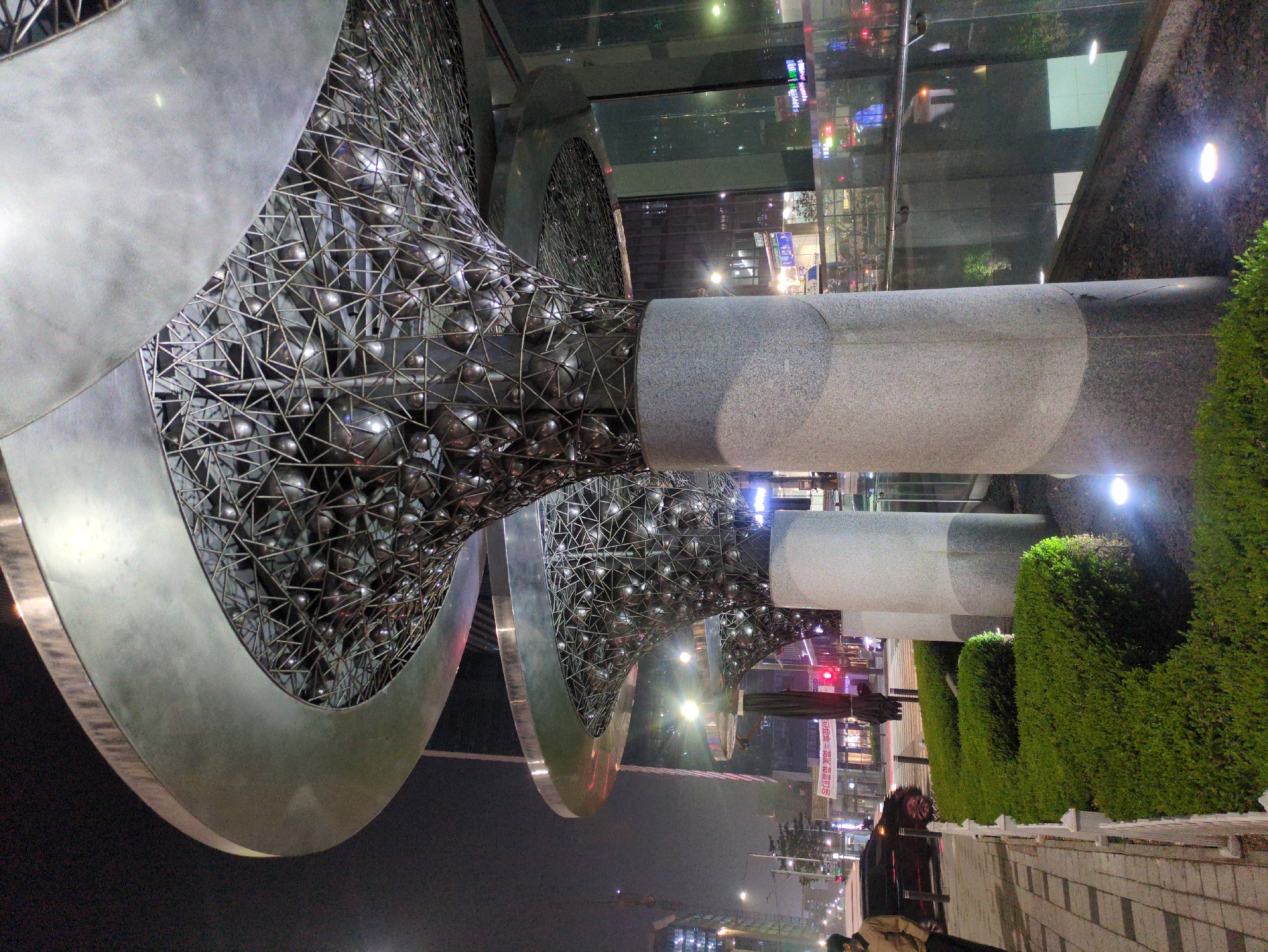 The image captures the sculpture "Twist Tower" in Seoul, South Korea, illuminated at night. The sculpture consists of two metal structures, each formed by a twisted, open framework of intersecting metal triangles enclosing numerous small metal spheres. The structures have rounded metal bases and are supported by cylindrical stone pillars. The backdrop features a glass-fronted building reflecting city lights and other buildings. The street scene in front of the sculpture includes a row of well-maintained green hedges, a paved sidewalk with people walking, and street lighting. In the distance, city lights stretch into the night sky. The overall setting is urban.