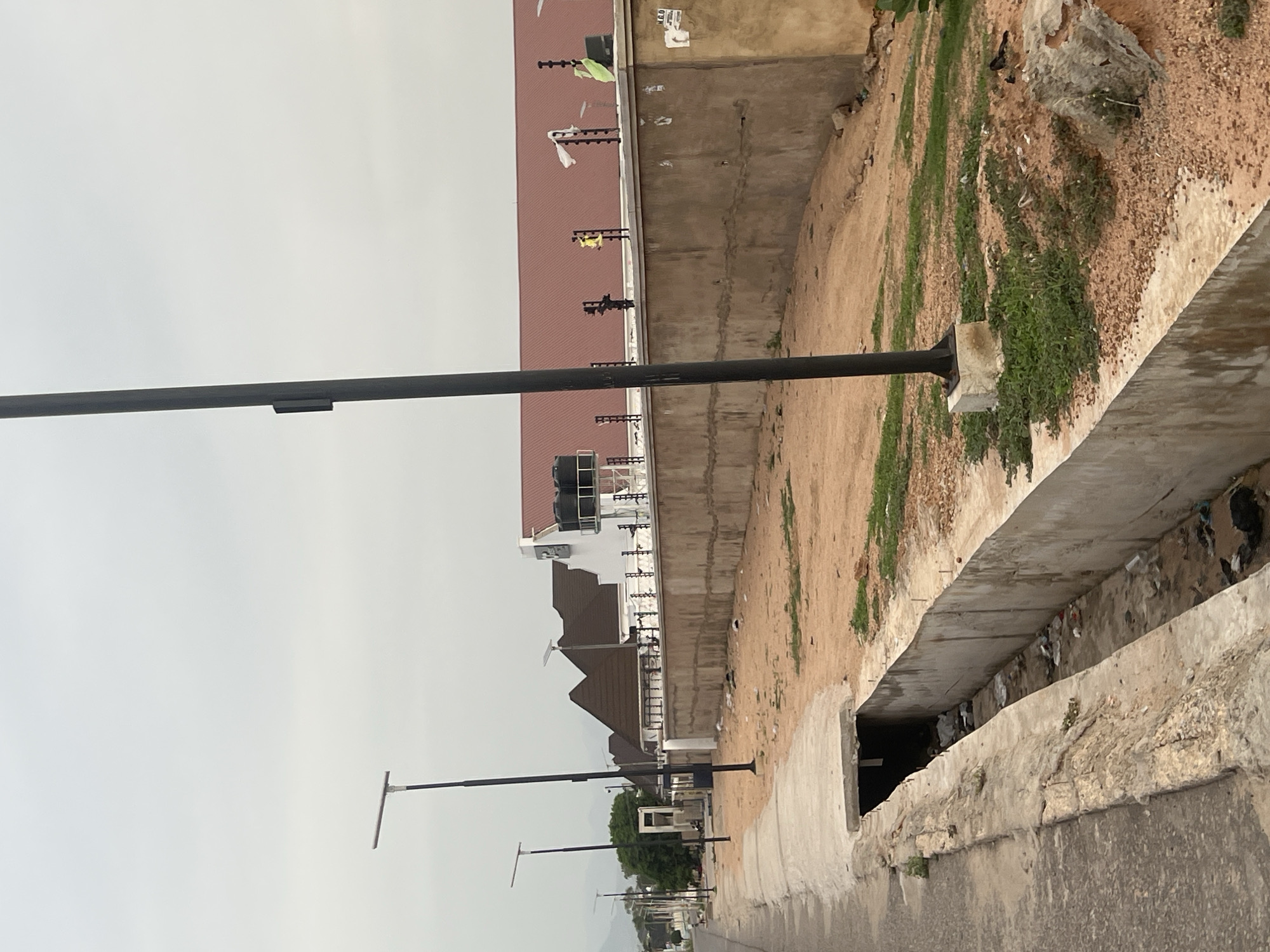 The image, taken in Jalingo, Nigeria, depicts an outdoor urban or semi-urban street scene under an overcast sky. The overall color palette is muted, dominated by shades of grey, brown, and the reddish-brown of a roof.
In the foreground, a tall, dark grey street light pole stands prominently on the right side of the frame, its base anchored in a light-colored concrete block. To its left, a concrete drainage ditch runs horizontally, partially open and revealing accumulated debris and litter within. A paved road surface is visible in the lower right corner, adjacent to the ditch.
Beyond the drainage ditch, an earthen embankment rises, covered in patches of dry, reddish-brown soil and sparse green grass. A grey concrete wall runs along the top of this embankment. Behind the wall, a building with a distinct reddish-brown roof and lighter-colored walls is visible. Along the top of the wall and parts of the building, there are dark, spiky security fence installations, with several pieces of light-colored fabric or plastic bags, and a green cloth, caught on their points.
Further in the distance, along what appears to be the same road, several more similar street light poles with horizontal light fixtures are visible, diminishing in size due to perspective. Another building with a dark, multi-peaked roof stands further back. The background is completed by a line of green trees against a uniformly light grey, overcast sky. There are no people visible, and no legible text can be discerned. The scene suggests a quiet, daytime moment in a developing urban landscape.