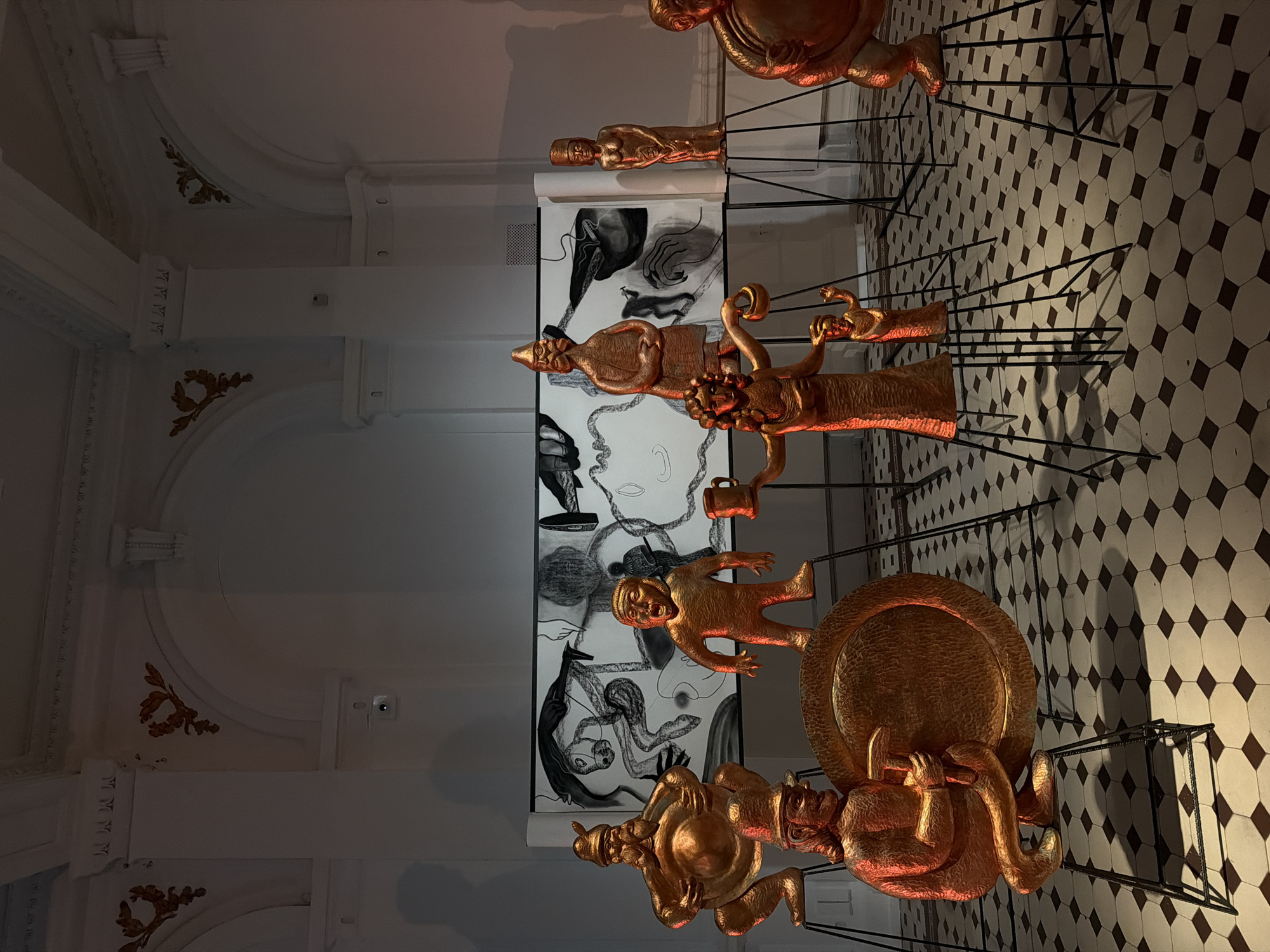 This media file captures an art installation or exhibition set within an ornate, possibly historical, interior space. The image, rotated 90 degrees counter-clockwise, displays a series of golden-colored, stylized figures arranged on black metal stands.
The figures depict human or human-like forms in various poses, some appearing grotesque or cartoonish. From left to right (when correctly oriented), there's a seated figure holding a large circular object, followed by a standing figure with a pointed hat, another figure with multiple limbs, a smaller child-like figure, and a larger, plump seated figure at the far end. The golden material of these sculptures reflects light with a warm, reddish-orange glow, creating strong shadows on the floor.
Behind the figures, large black and white drawings or prints are mounted vertically. These artworks feature surreal imagery, including distorted faces, hands, abstract shapes, and elongated figures, rendered in a illustrative, almost dreamlike style.
The setting itself is notable for its architectural details. The ceiling and upper walls are white with intricate molding and decorative gold-leaf accents, suggesting a classical or baroque influence. Visible structural beams are also painted white. The floor is covered in a distinctive black and white octagonal tile pattern, further contributing to the historical or grand aesthetic of the room. The lighting appears to be artificial, with spotlights illuminating the sculptures and casting dramatic shadows.
No visible text or specific identifying marks are present on the artworks or within the scene. The overall impression is one of a curated artistic display in an elegant, somewhat dimly lit environment.