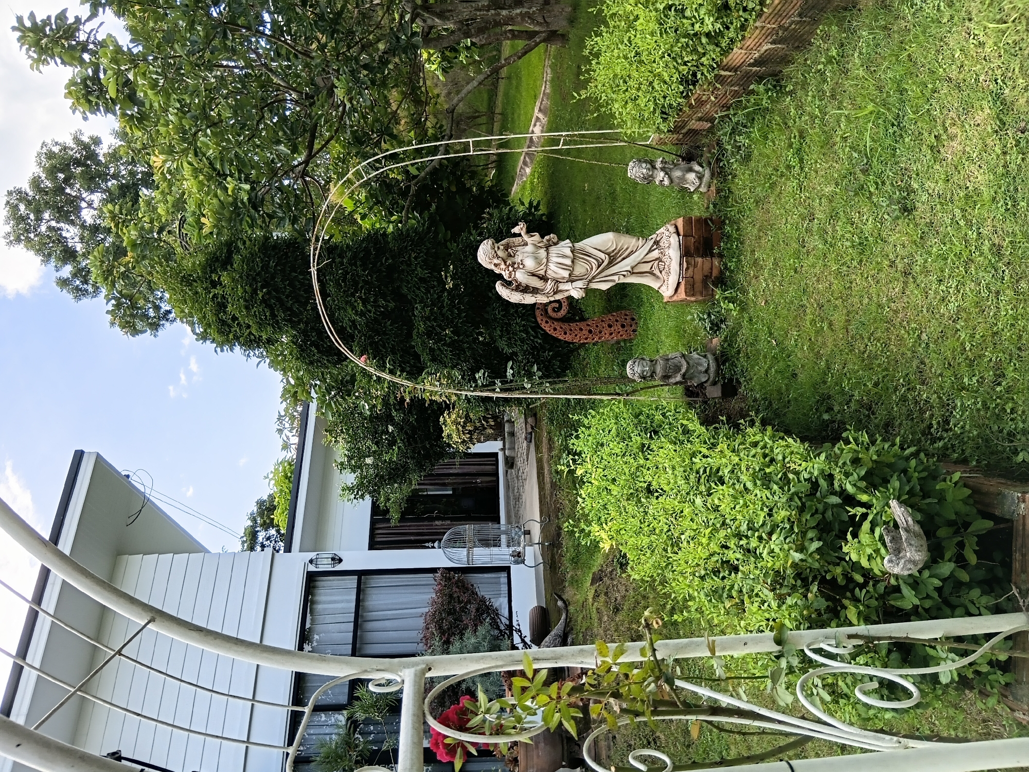 The image depicts a garden scene in Ban Tong, Thailand, featuring a white arched structure in the foreground with a lush green plant growing on its left side and a red rose in bloom. Beyond the arch, a white statue of an angel stands prominently on a brick pedestal, holding a small bird in its hand. Flanking the angel statue are two smaller grey statues of cherubs or children. The garden is well-maintained with neatly trimmed bushes and a grassy lawn. In the background, a white building with a dark-trimmed roof and windows is visible, along with mature trees providing shade. The sky is partly cloudy. The scene evokes a sense of tranquility and artistic decoration.