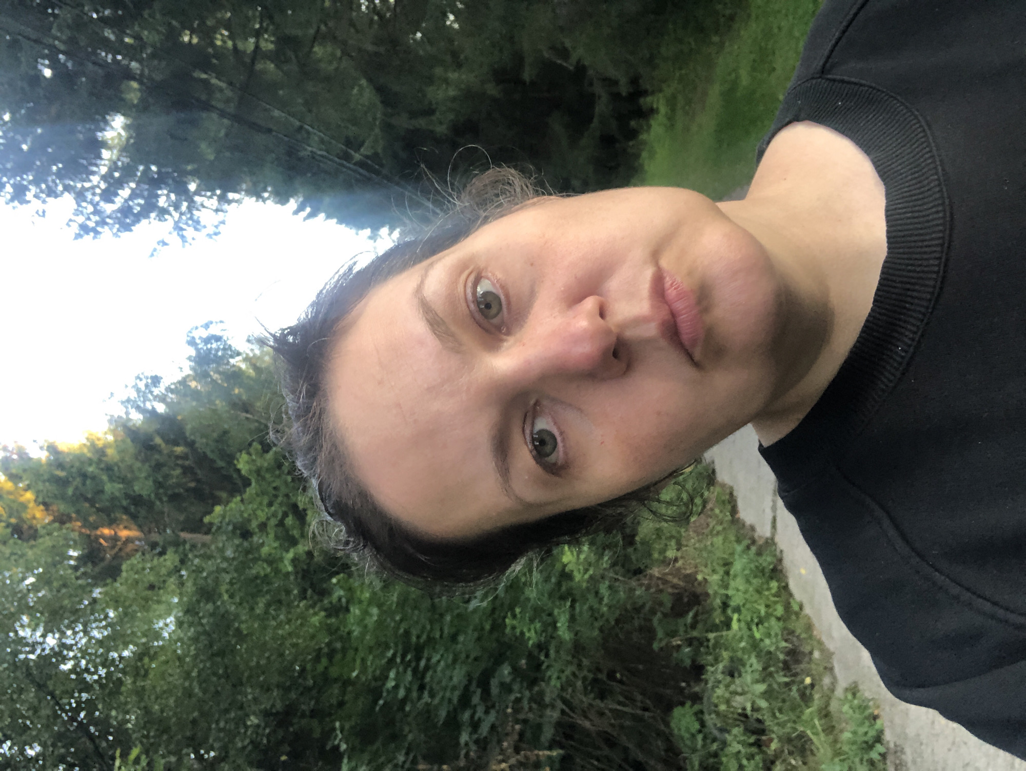 A woman of East European descent, appearing to be in her 40s, stands on a paved path in a wooded area. She is wearing a black sweatshirt. Her hair is dark and pulled back. Her expression is neutral, with her head tilted slightly to the right. The background consists of dense green foliage and tall trees, with a bright sky visible through the branches. The paved path extends into the distance, bordered by greenery. The lighting suggests it is daytime, possibly late afternoon, with sunlight filtering through the trees. The specific location is Granat, Russia.