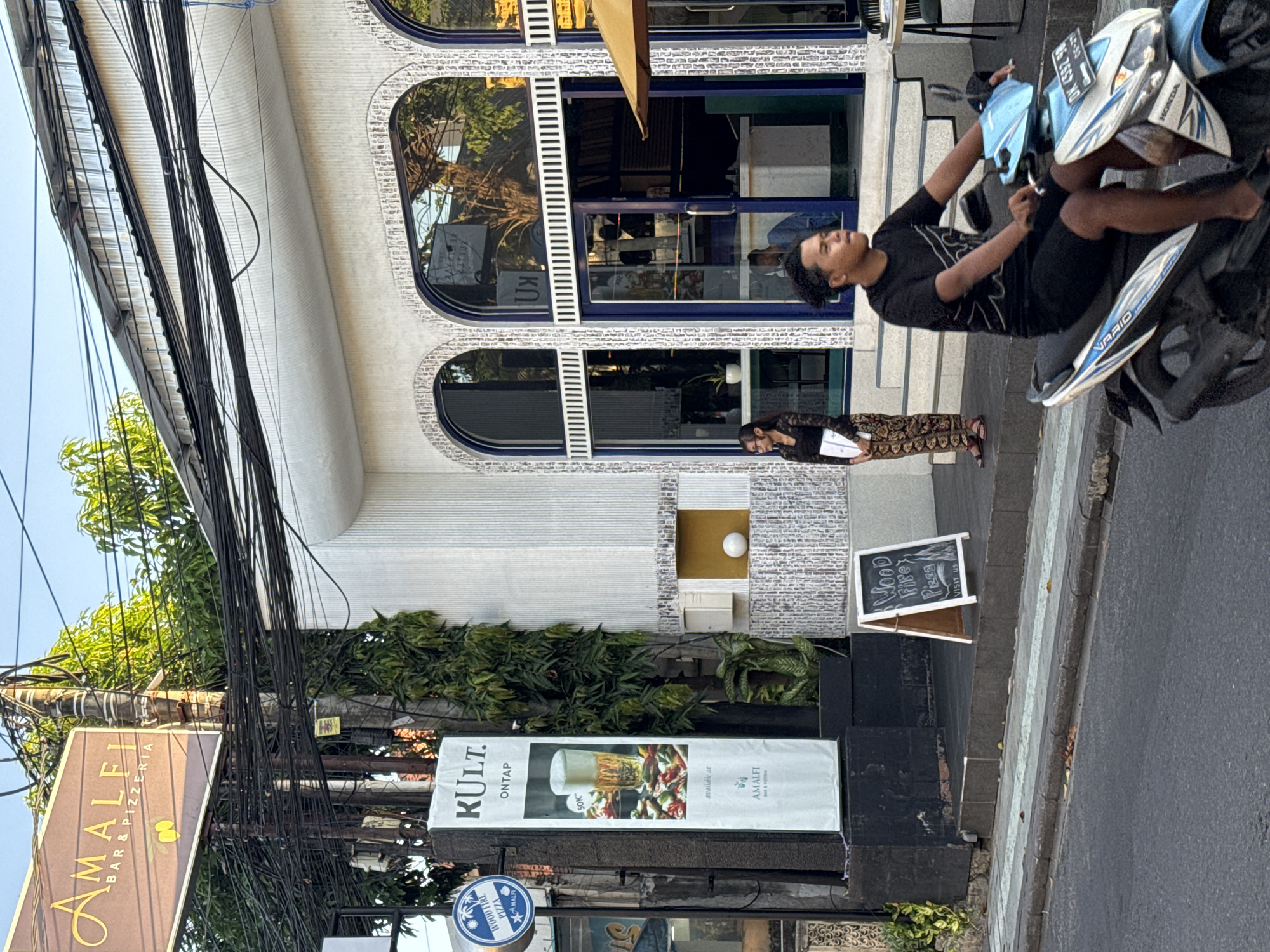 In Kuta, Indonesia, on a street, a man is riding a motorcycle. In front of a building, a woman stands next to a poster. The building has arched windows and a textured white facade. A sign for "AMALFI BAR & PIZZERIA" is visible. The scene is captured in daylight.