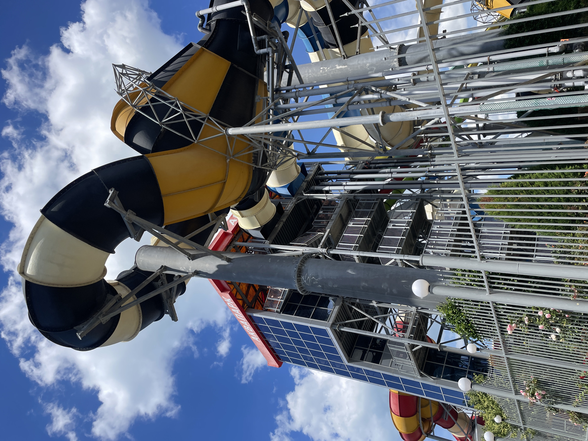 An exterior, low-angle shot captures a section of a water park with a prominent, multi-colored waterslide structure against a bright, partly cloudy sky. The main slide sections visible are wide tubes, painted in alternating black and off-white stripes, and yellow and black checkered patterns. These tubes twist and curve, supported by a complex network of grey metal scaffolding and pipes.
To the left, a tall, rectangular building with a red roof and blue-tinted glass windows rises vertically. Text on the red overhang reads "АКВАПАРК" in white letters. On the left side of this building, a red and yellow striped waterslide tube is partially visible. Metal walkways with railings ascend the side of the building, with people visible on some of the platforms.
In the foreground, a grey metal fence with vertical bars runs horizontally across the image. White, spherical lights are mounted on posts along the fence. Pink roses and green foliage are visible behind the fence, adding a touch of nature to the scene. The overall impression is one of a vibrant, active water park on a sunny day.