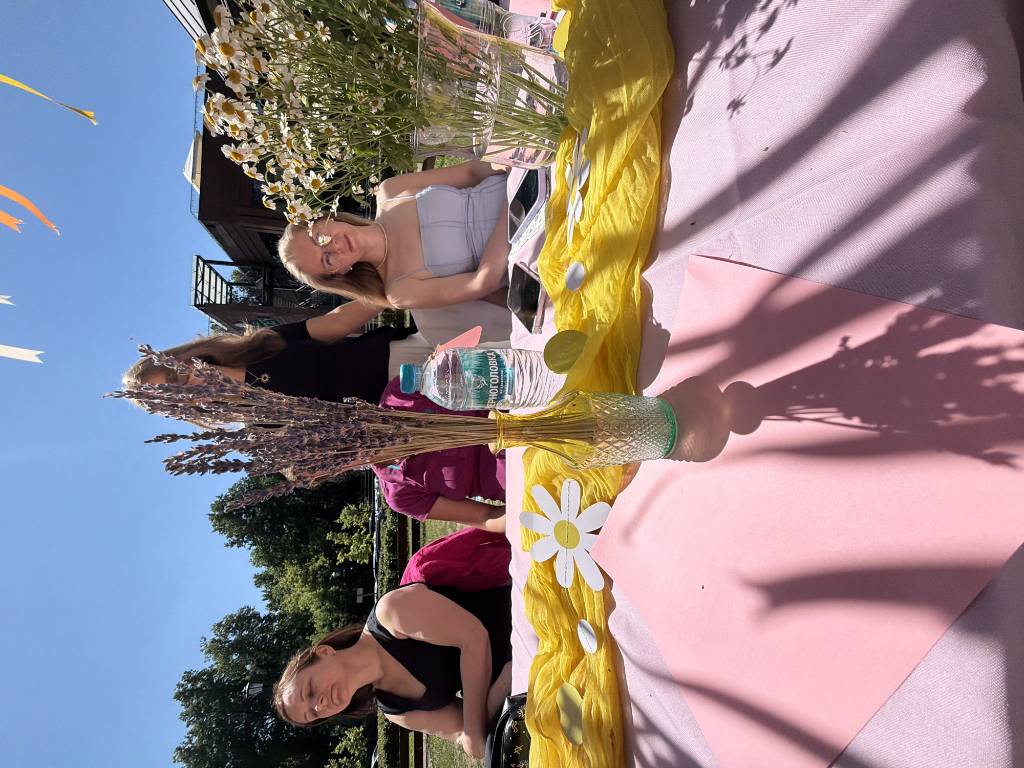 In Vidnoye, Russia, on a sunny day, two women are seated at a table. The woman on the left, with her eyes closed, wears a black sleeveless dress and sits behind a pink tablecloth. A pink jacket is draped over her chair. To her right, a woman in a light blue dress with thin straps looks towards the camera, wearing glasses and a necklace. She is seated behind the same table. Between them, a bouquet of dried lavender is in a patterned amber glass vase, and a bottle of water stands nearby. The table is decorated with a bright yellow fabric and a paper daisy. In the background, a building with a modern design and trees under a clear blue sky are visible.