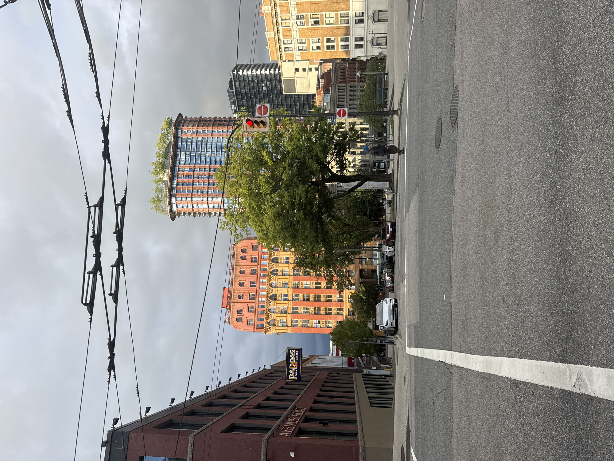 The image depicts a street scene in Vancouver, Canada, with a focus on an intersection under an overcast sky. In the foreground, a road with white lane markings runs across the frame, leading towards several vehicles stopped at a traffic light. To the left, a modern brick building with large windows is visible, featuring a sign that reads "AVENUE ROAD." Further down the street, a vintage-style orange and yellow building with arched windows stands out. Behind it, a contemporary tall building with a green rooftop garden rises into the sky, contrasted by other high-rise structures in the background. A lone pedestrian, wearing a blue hat and dark clothing, is crossing the street. Overhead, trolley bus wires crisscross the sky, indicating public transportation infrastructure. The overall atmosphere is that of a typical urban environment.
