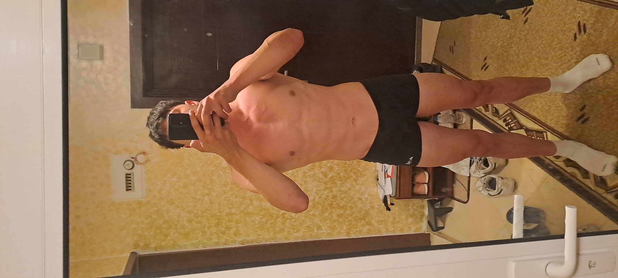 A man is taking a mirror selfie in what appears to be a bathroom or bedroom. The man is wearing black swim briefs and white socks. His body is visible from the head down. He is holding a black smartphone with both hands over his face. The reflection in the mirror shows a door, patterned wallpaper, and a switch box. The floor is covered in a patterned rug, and there are various objects including a pair of shoes, a water bottle, and a small cabinet. There is no information available in the picture about the location in the city of Burgas, Bulgaria.